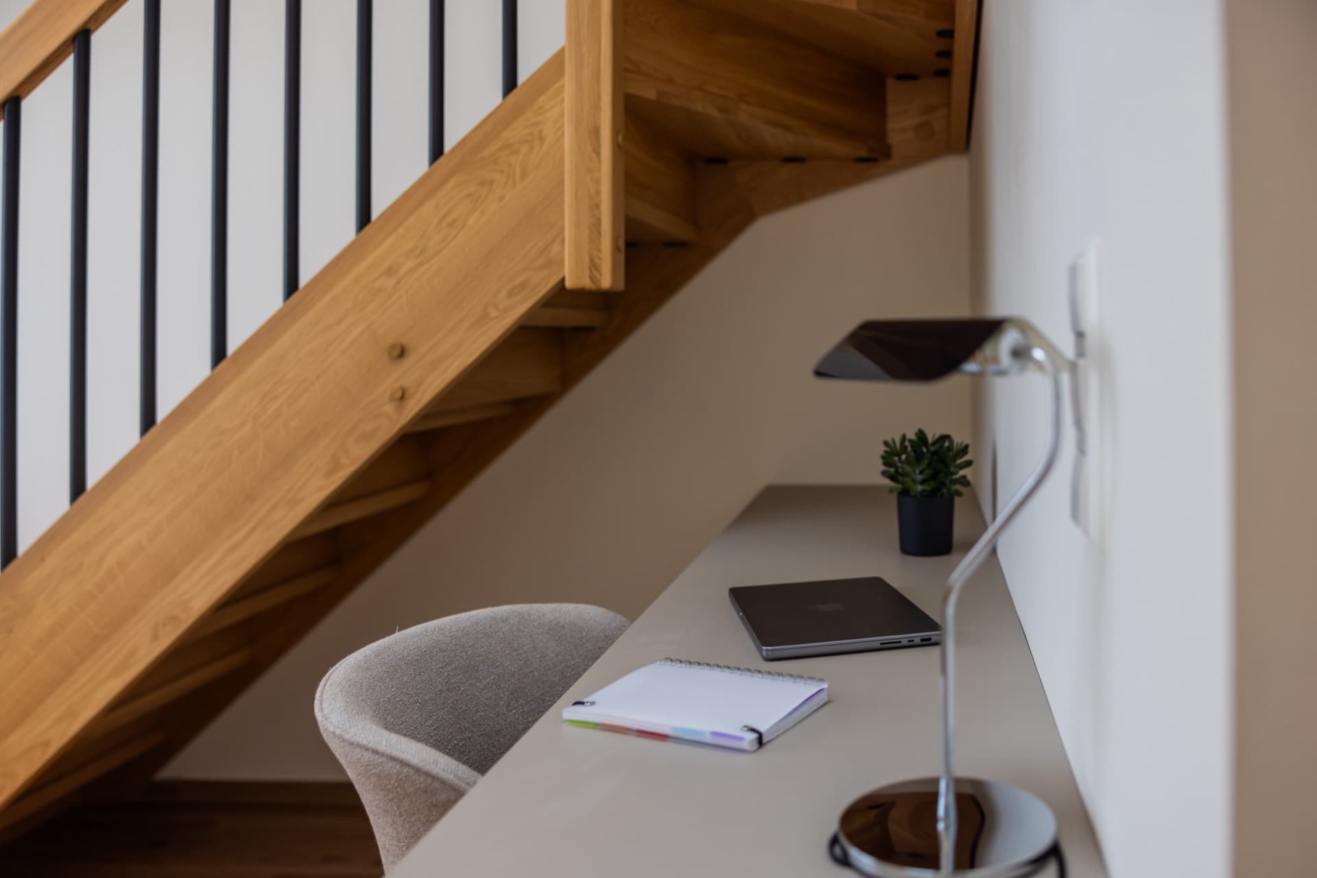 Close-up image of the working area of the bedroom for rent. It depicts a working area with a desk, and a comfortable chair. On the desk, there is a laptop and a lamp.
In the background wooden stairs leading to the bedroom can be seen