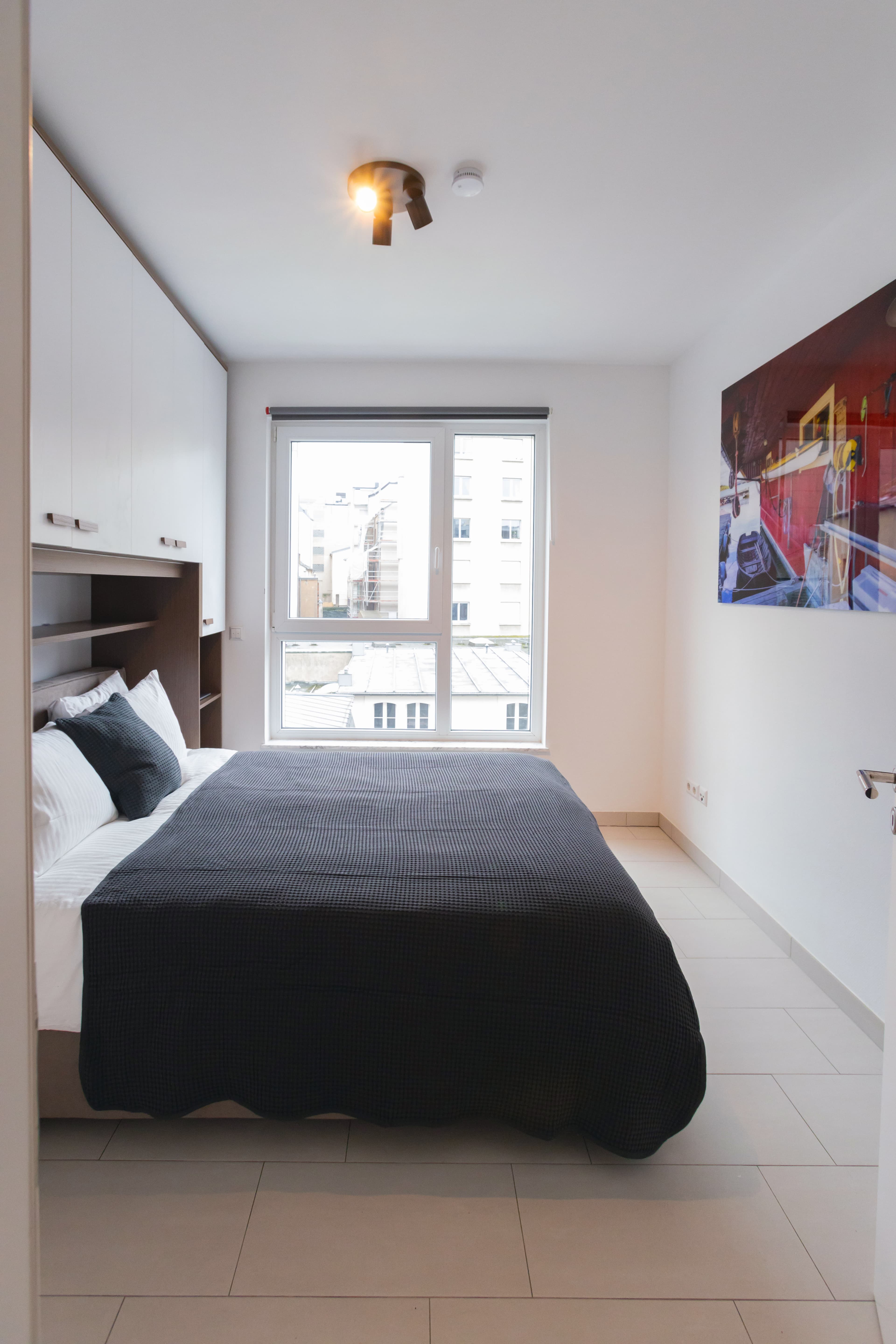 Bedroom with a large bed with sheets and cushions. The bed is combined with measured dressings. On its right, there is a floor-to-ceiling window, and in front of it, a big picture hangs on the wall.