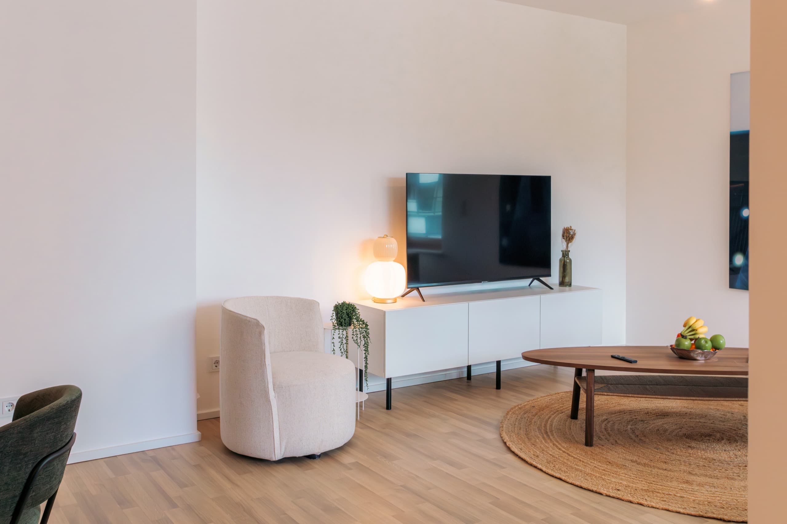 Overview of the lounge area in Apartment Escher, Media Bay Apartments, Kirchberg, Luxembourg city. A white TV stand with a big LCD television, a white armchair, and a coffee table on a carpet.