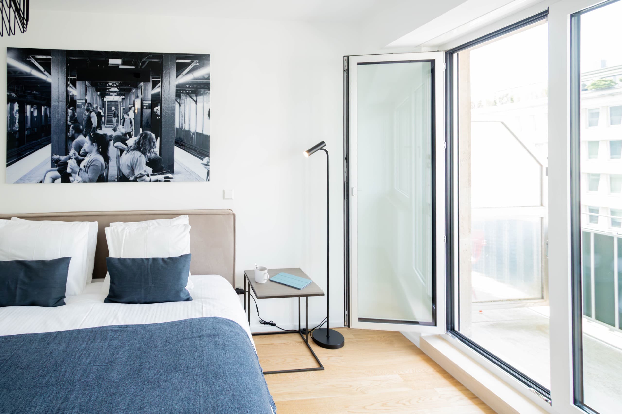Bedroom with a large bed with white sheets and pillows. A nightstand and a standing lamp on its left. Open balcony floor-to-ceiling door next to it.