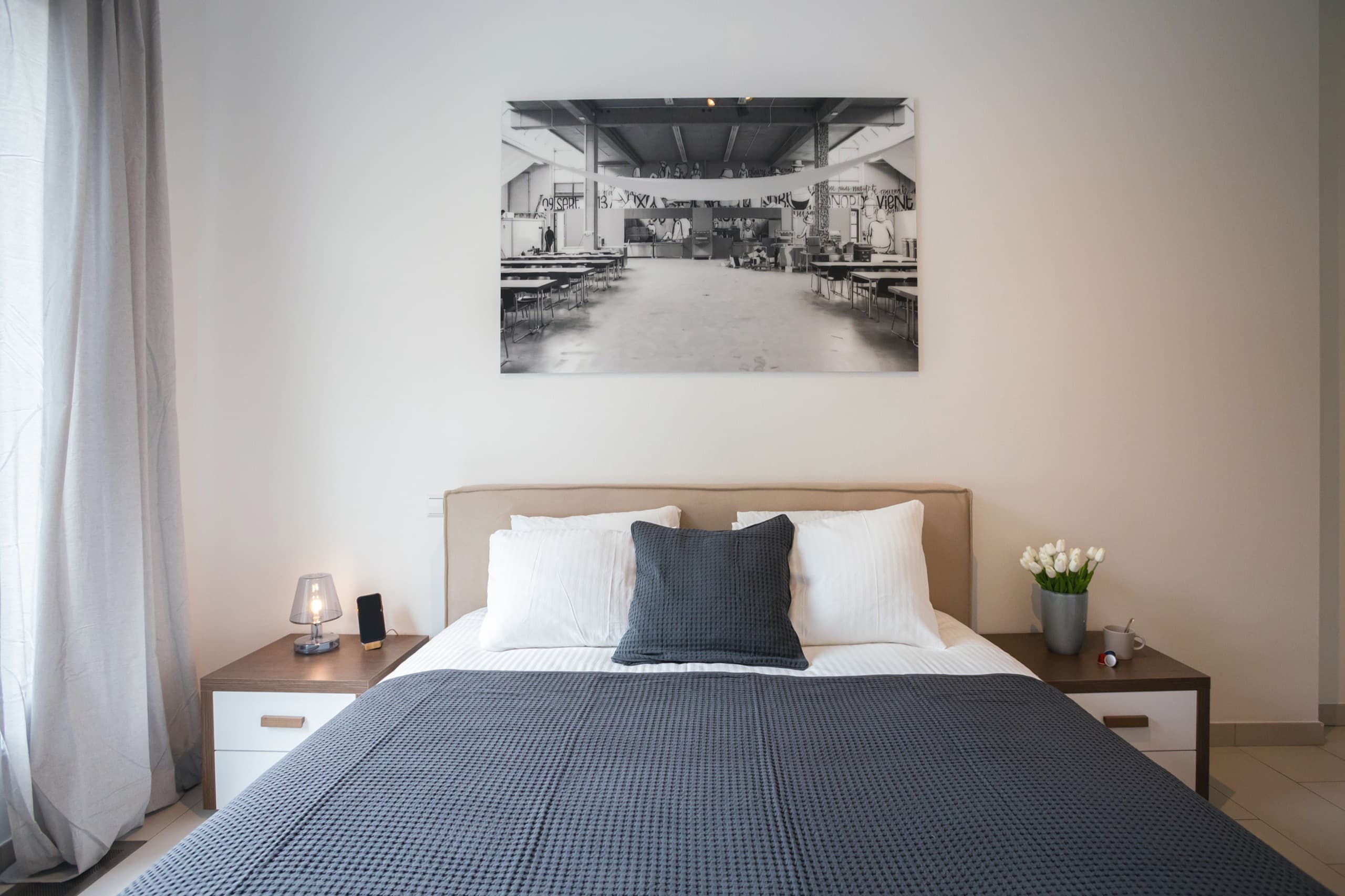 Bedroom with a large bed with white sheets, pillows, and a dark blue cover. The bed is flanked by two nightstands, one with a wireless phone charger on it. On the wall, above the bed hangs a big picture. On the right, there is a window with curtains.