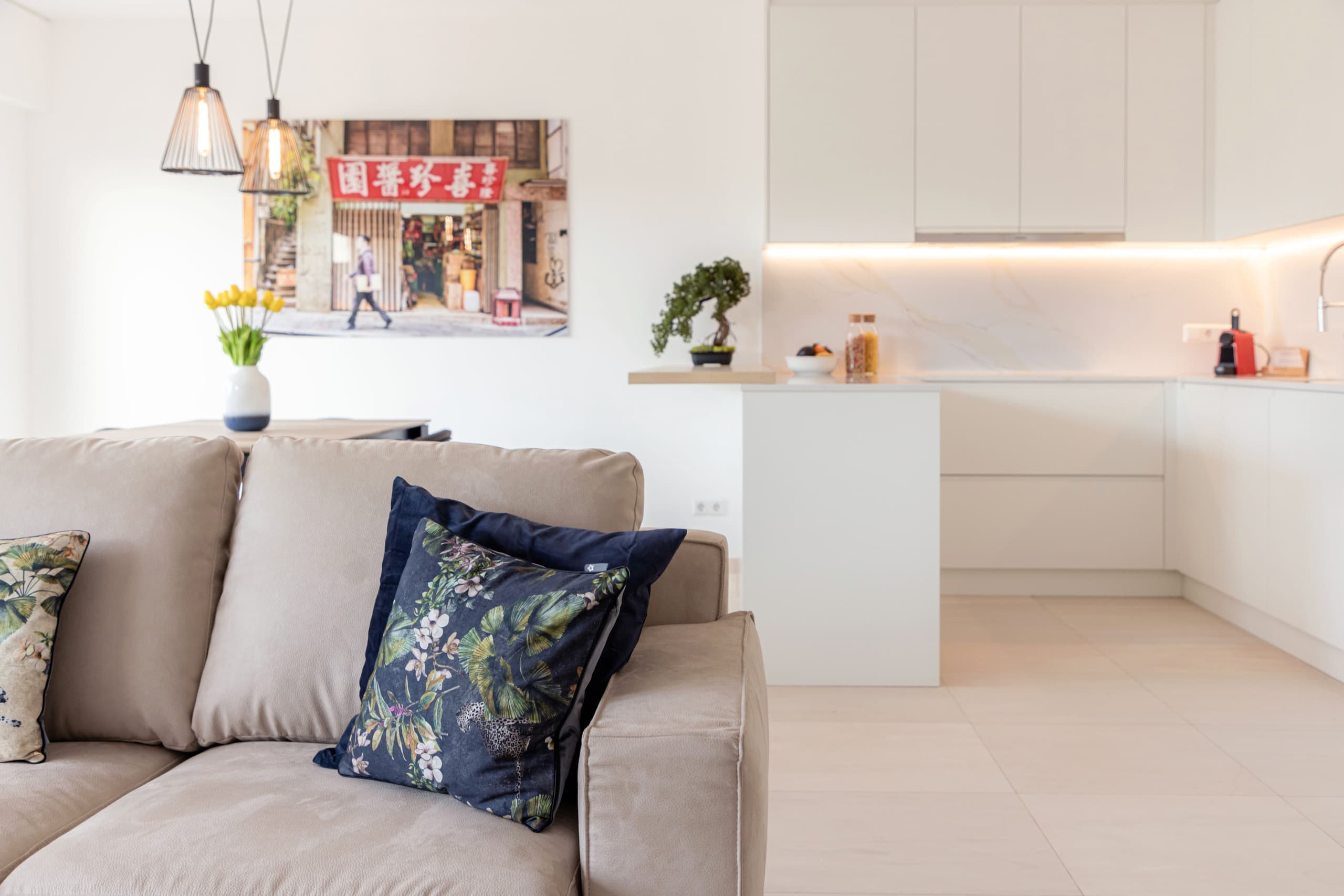 In the background, a table with a flower vase on it, and a picture of pedestrians hanging on the wall. Next, on the right, is the kitchen with a bonsai flower pot on a counter. In the foreground, is a beige couch with multi-color cushions on it.