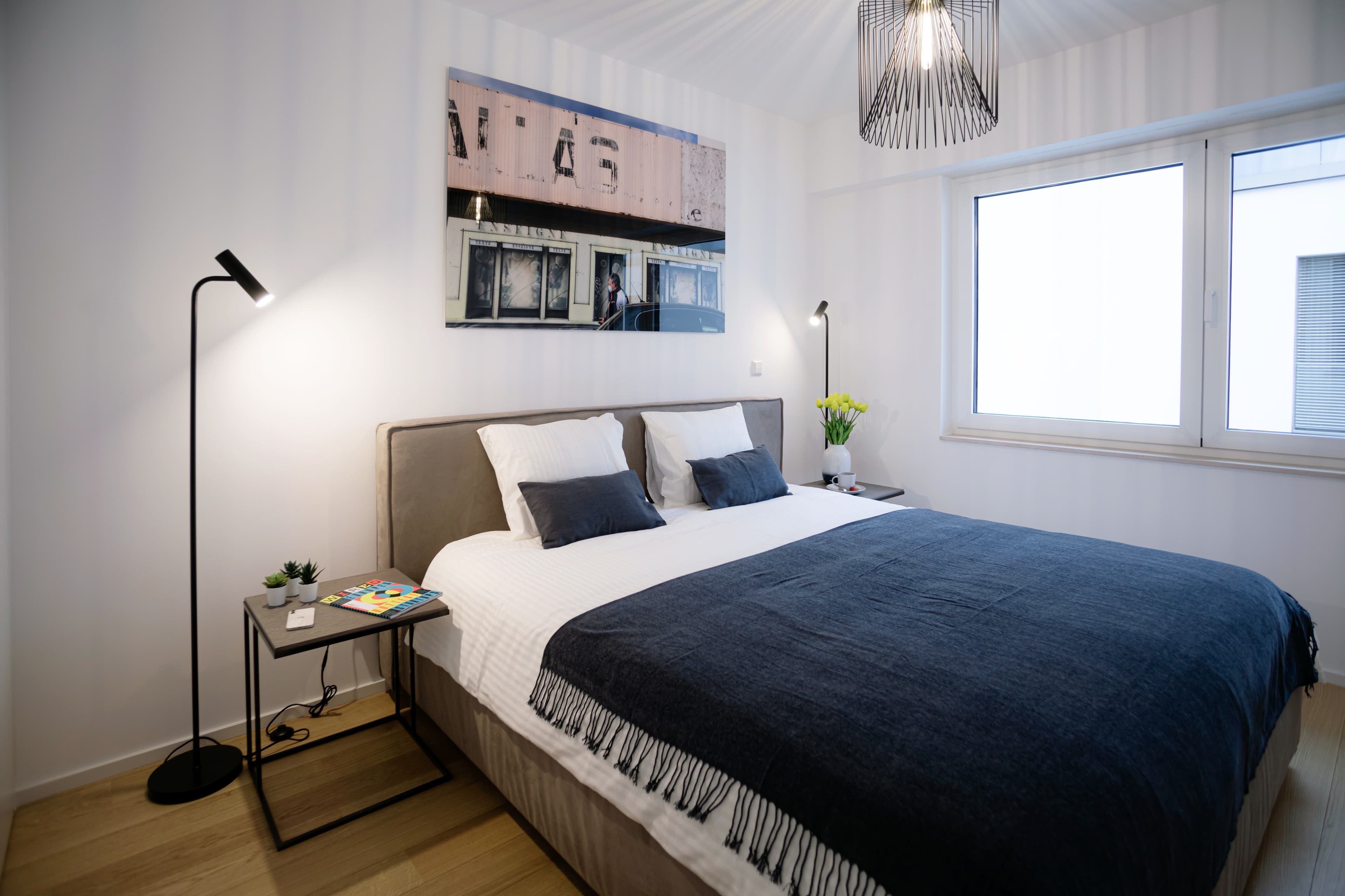 Bedroom with a large bed with white sheets and dark blue cover. The bed is flanked by two lamps and nightstands. Behind the bed hangs a big picture with a street, and, on its right, there is a window.
