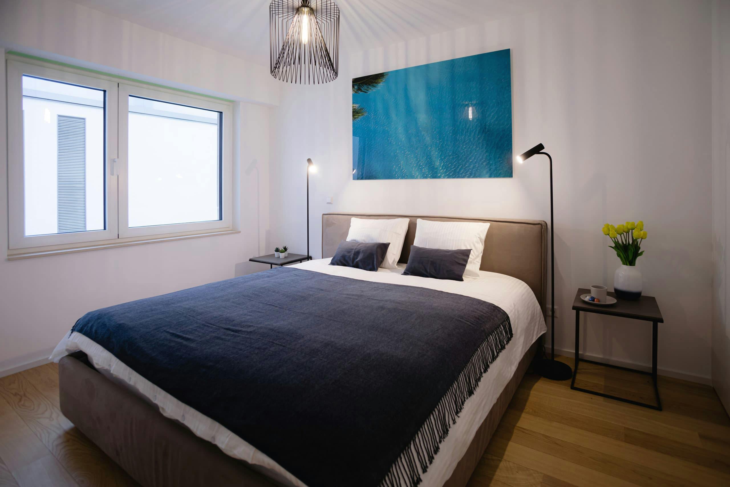 Bed with white sheets and blue cover, flanked by two lamps and nightstands. A blue sea picture hangs on the wall, behind the bed. On its right, there's a window.