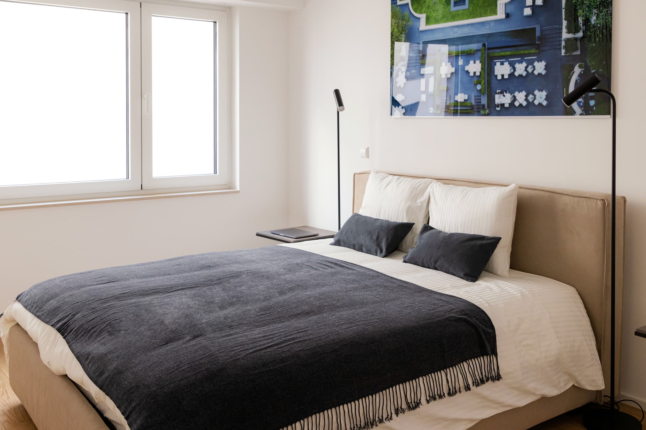 Bedroom with a large bed with white sheets and a dark blue blanket. The bed is flanked by two lamps and two nightstands. Above the bed, on the wall, hangs a big picture.
On the left side of the bed, there are two windows.
