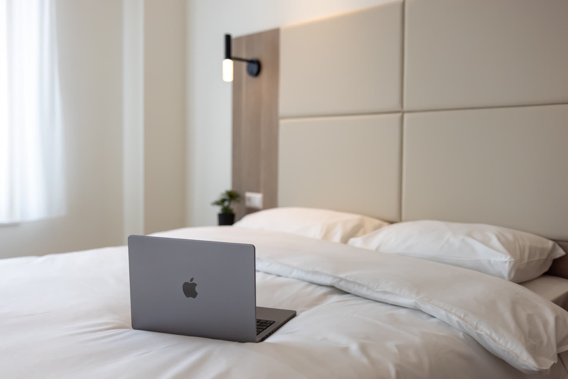 Close-up image of the bedroom's king-size bed, with a laptop on top. Next to the bed, a nightstand with a vase and a large window can be seen.