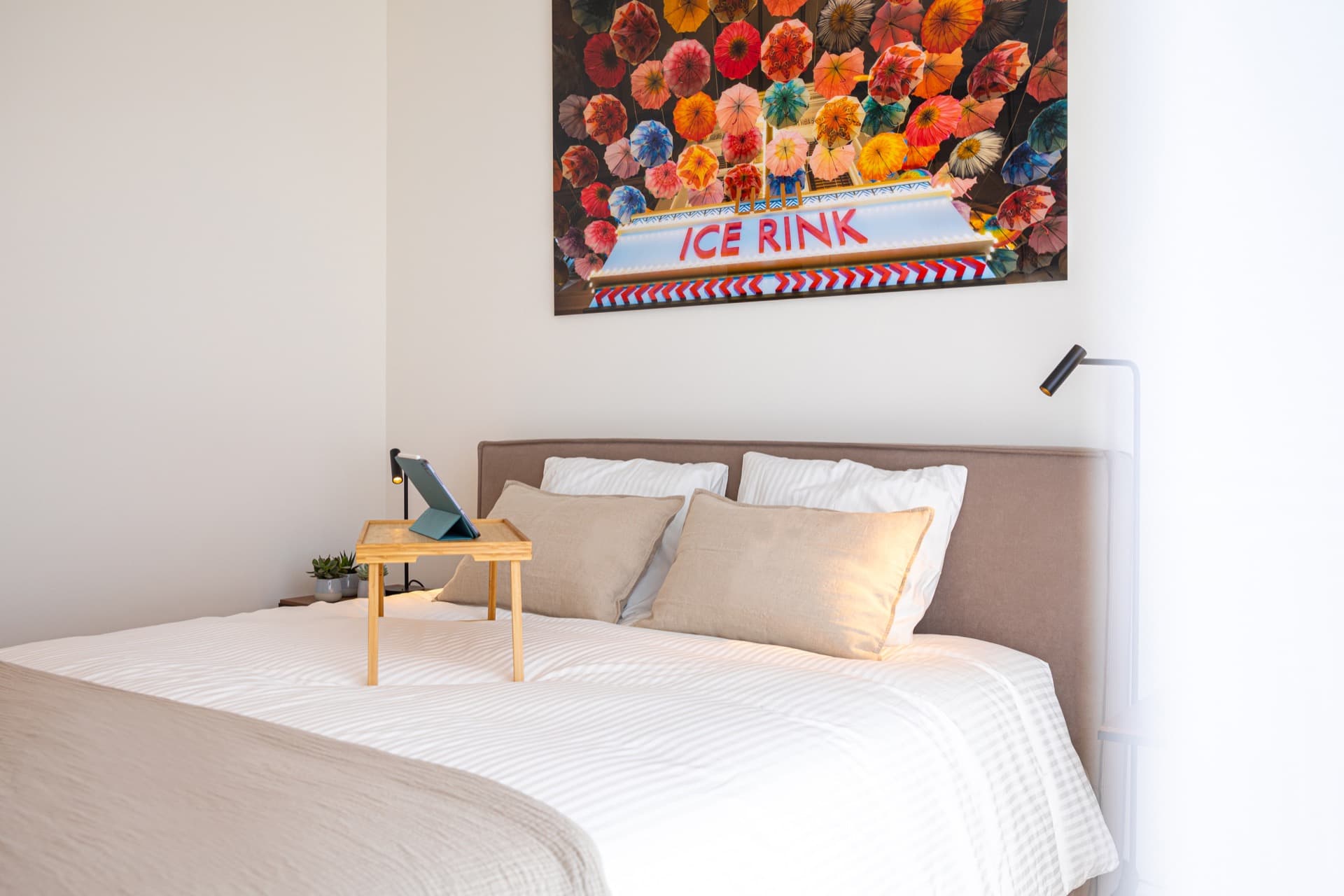 Bedroom with a large bed with tray and iPad, a bedside table with small vase and plants. Above the bed, a large photo of flowers on the wall.