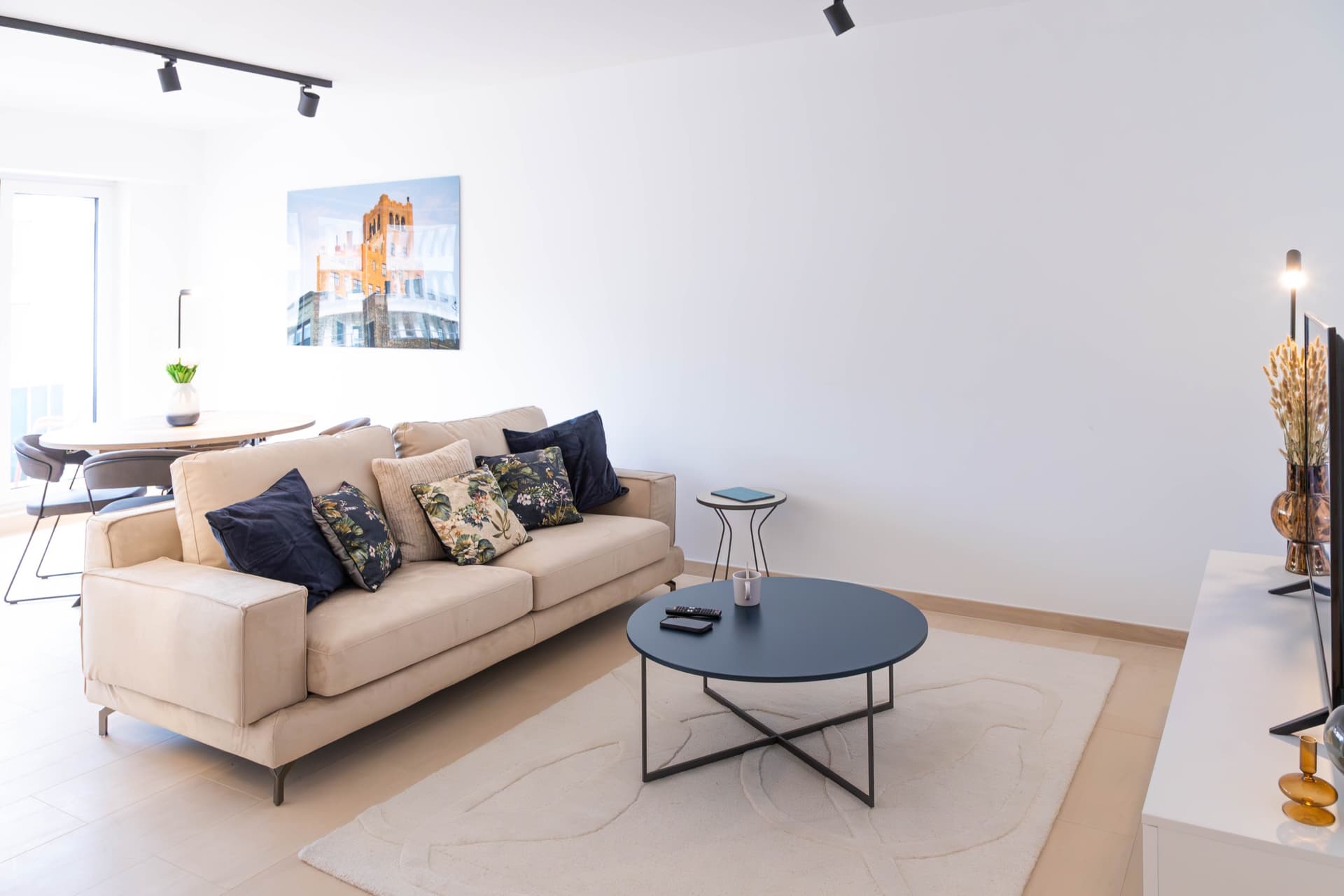 Lounge area with sofa and cushions. In front, coffee table and a white carpet underneath it. In front, a TV stand with a smart TV on it. Behind, a coffee table with a flower vase on top and floor-to-ceiling windows.