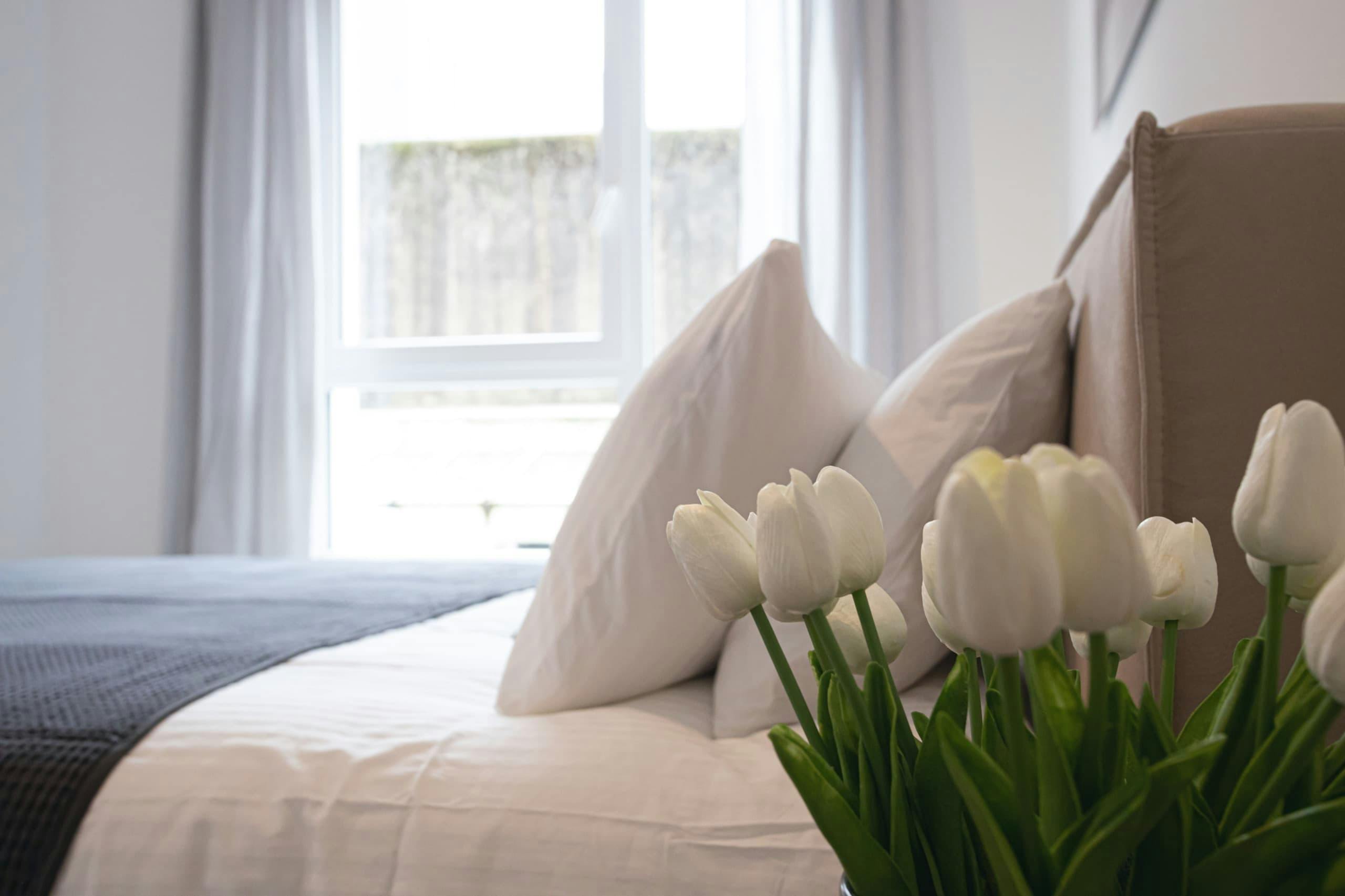 Close-up image with the bed, fluffy white pillows, and white tulips in a vase on a nightstand. Light comes through the big window.