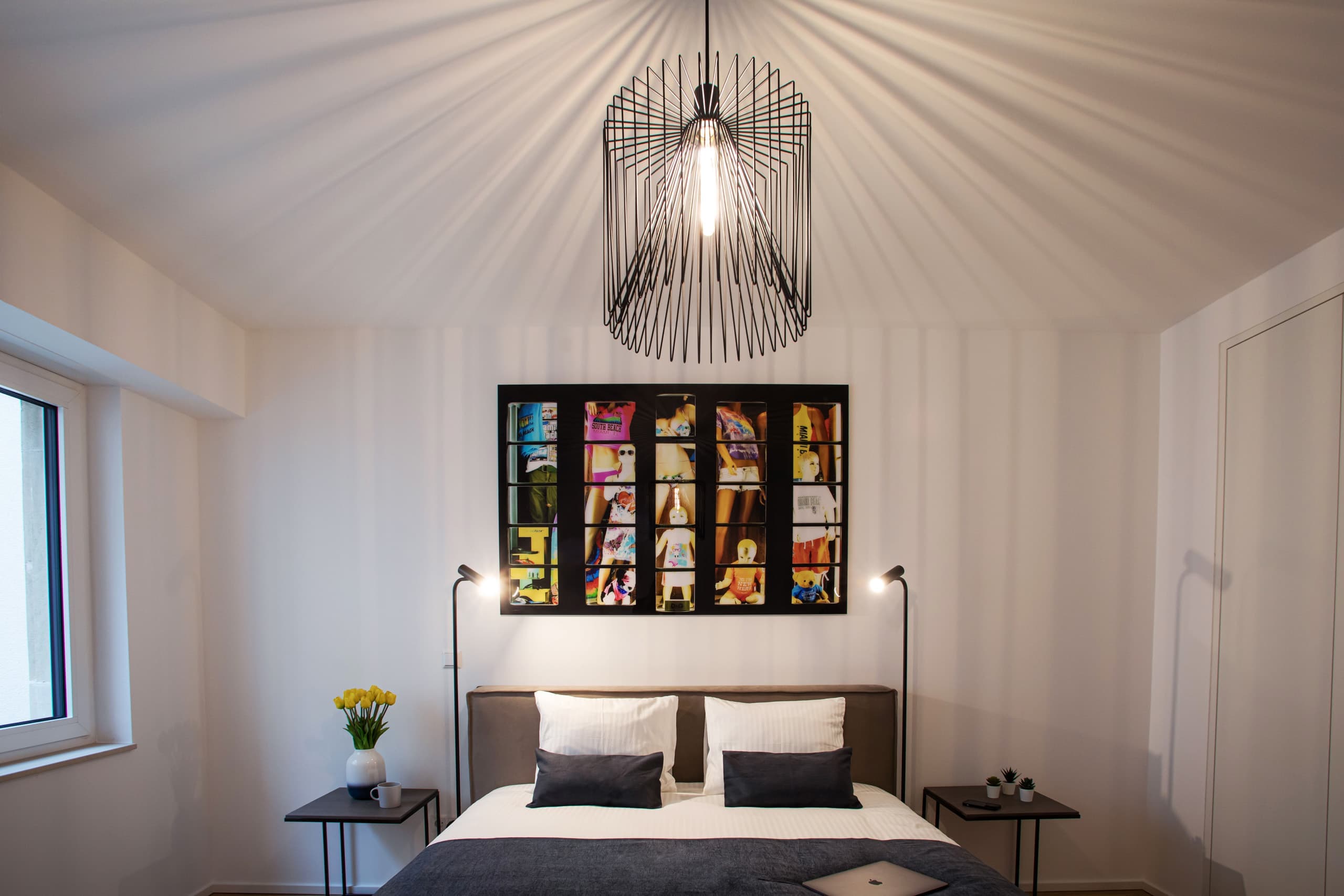 Second bedroom with a large bed with white sheets and a dark blue cover. The bed is flanked by two lamps and nightstands. On the left, there is the window.