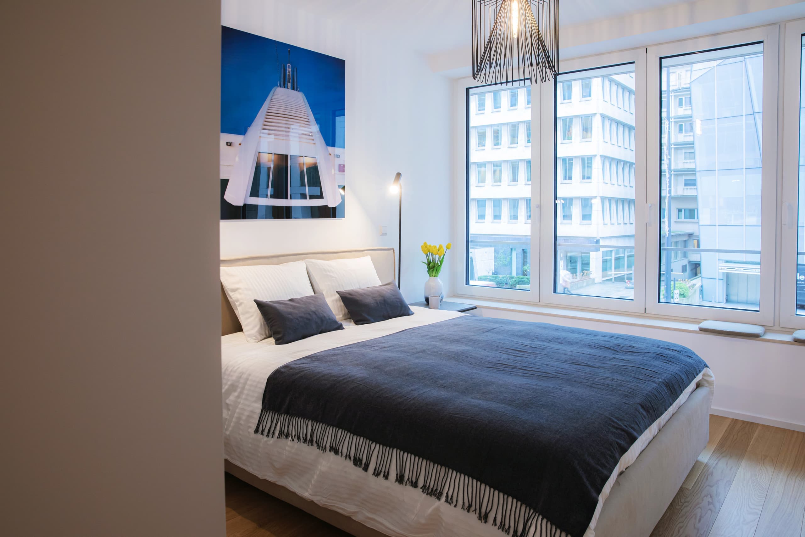 Bedroom with a large bed with white sheets and a dark blue cover. On the left, you can see big windows and a flower vase with yellow tulips in it.