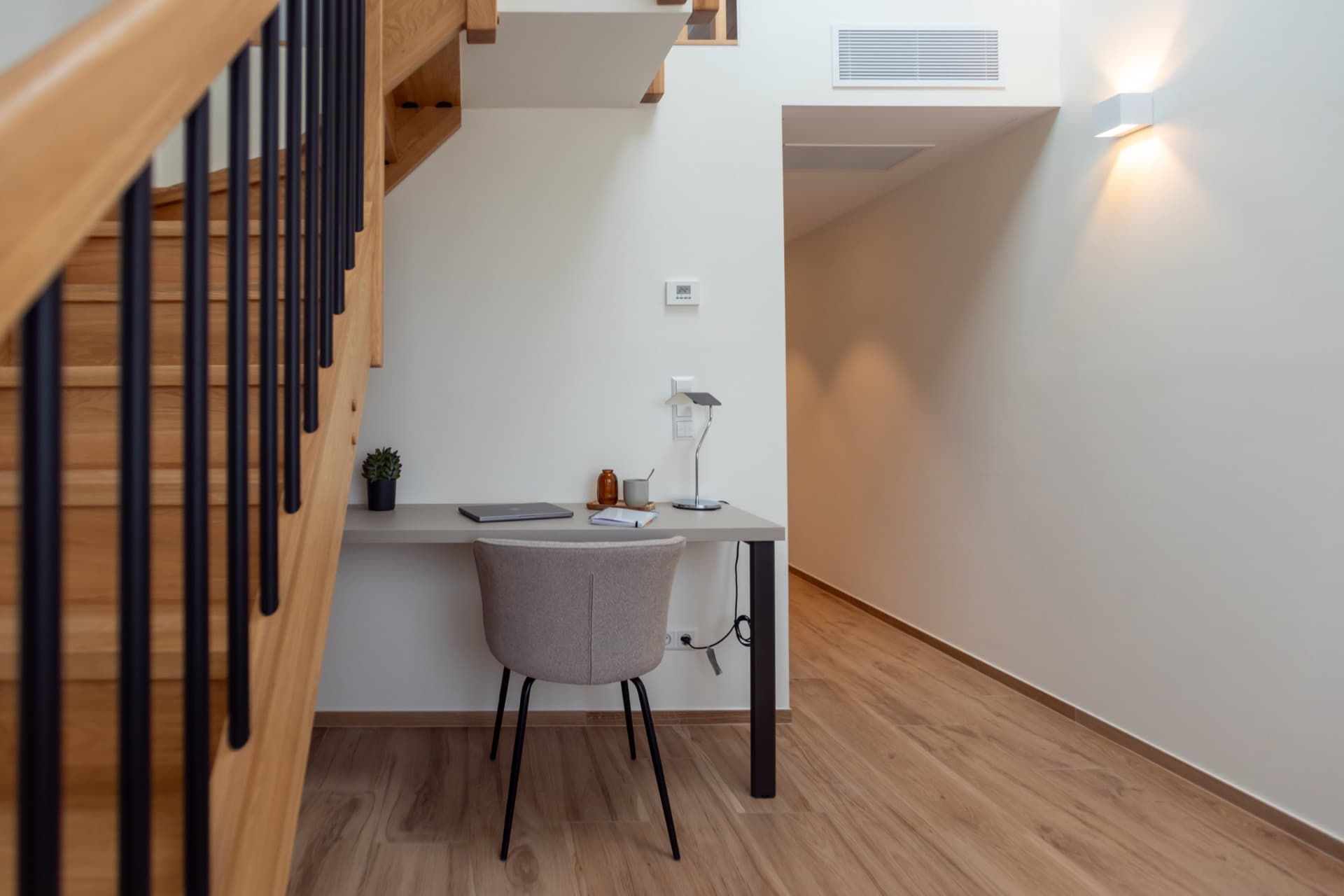 The picture depicts the working area in Mezzanine room to rent in Cessange House. It has a long hallway, a desk, a comfortable chair and stairs leading to the bed.