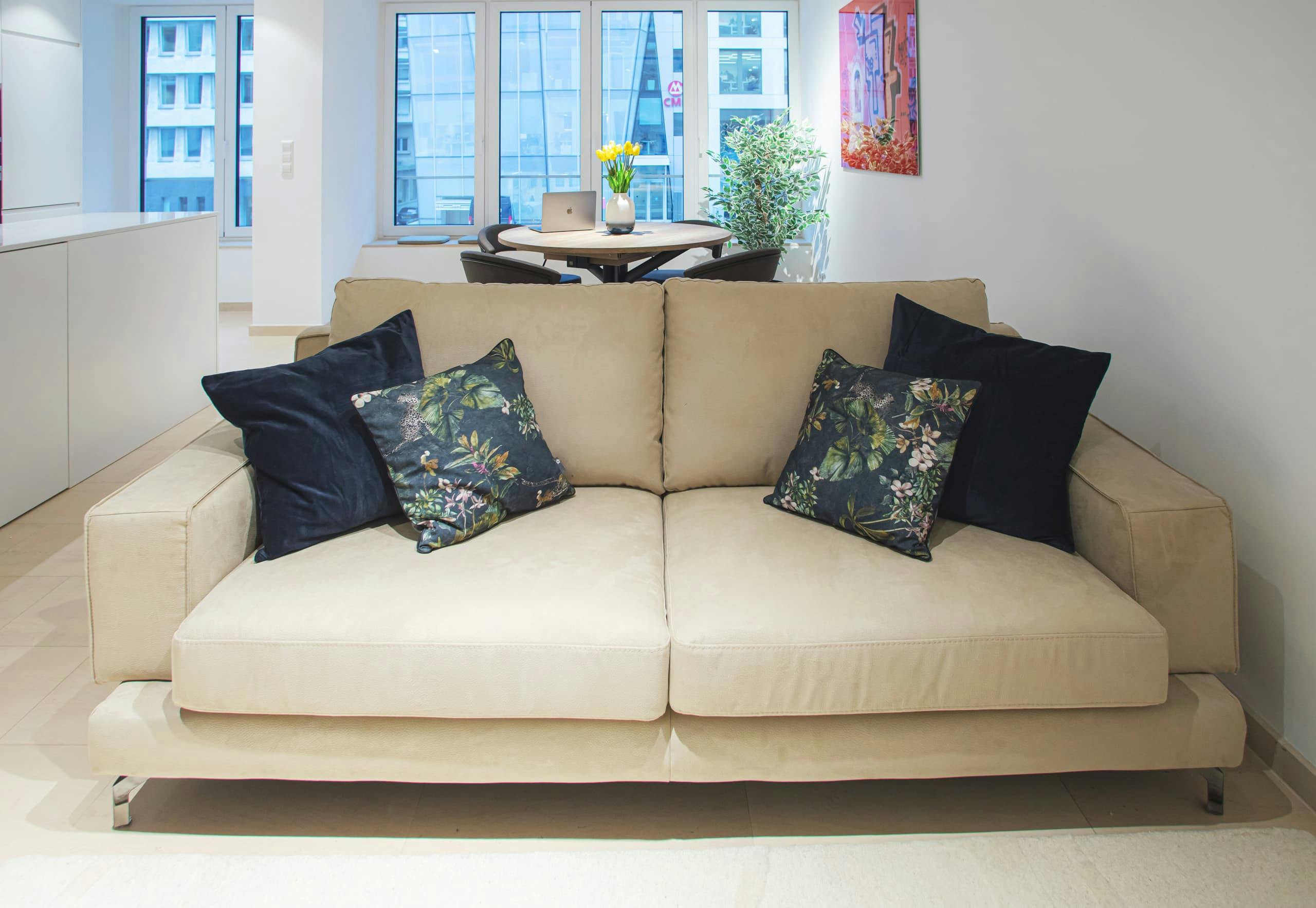in the foreground is a beige sofa with cushions on it. Behind it, in the background, is a dining table with chairs and big windows. On the left the corner of the countertop is visible, and on the right, a big picture hangs on the wall.