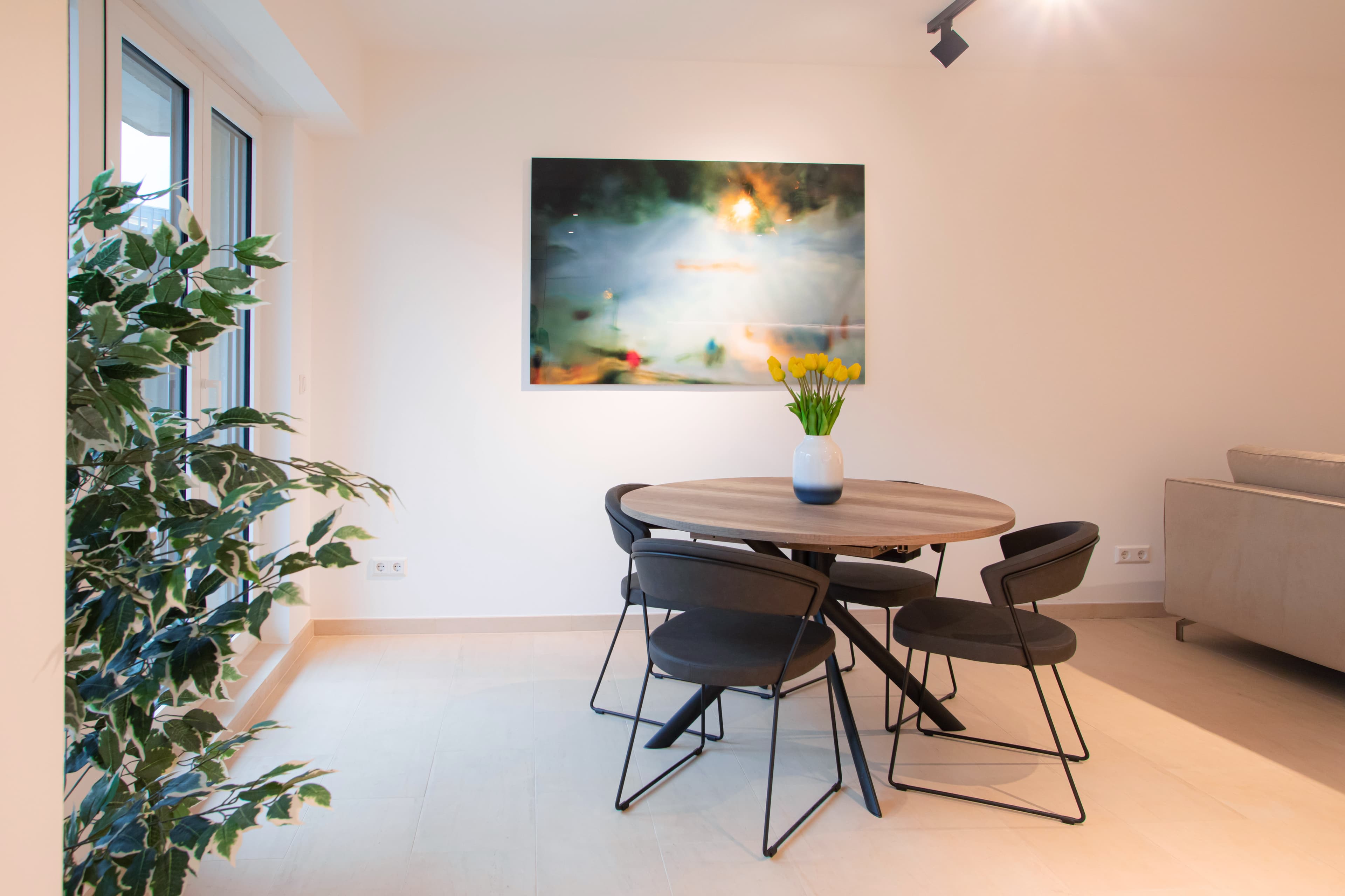 Dining table with a white vase with yellow tulips in it. on the left, large windows, and a big flower on a pot. On the wall hangs a big picture. On the right can be seen the back of the sofa.