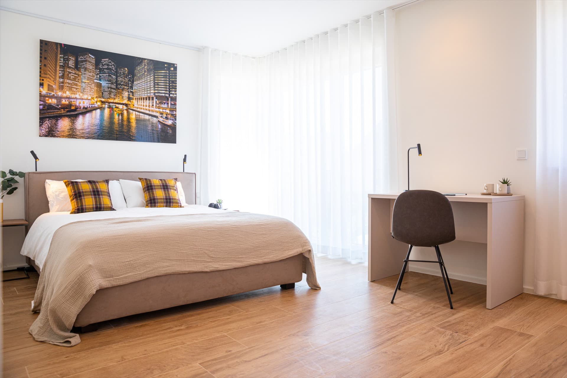 The picture depicts a bedroom with a bed with white sheets and cushions, a desk with a lamp and chair, and a large window.  Above the bed, there is a night-time marina picture.