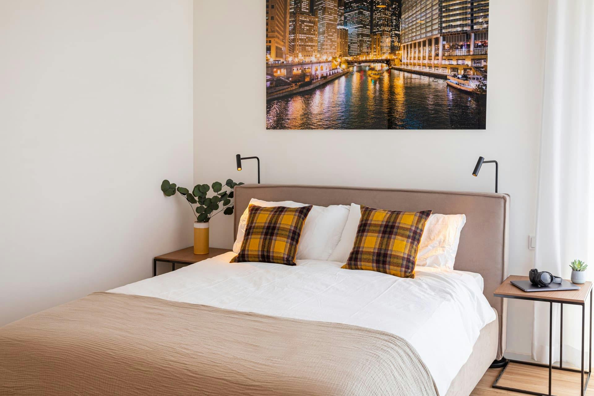 Picture of bed with two lamps and bedside table holding a vase of green foliage. White bedding and mustard tartan cushions on the bed, with a night cityscape photograph hanging on the wall. 