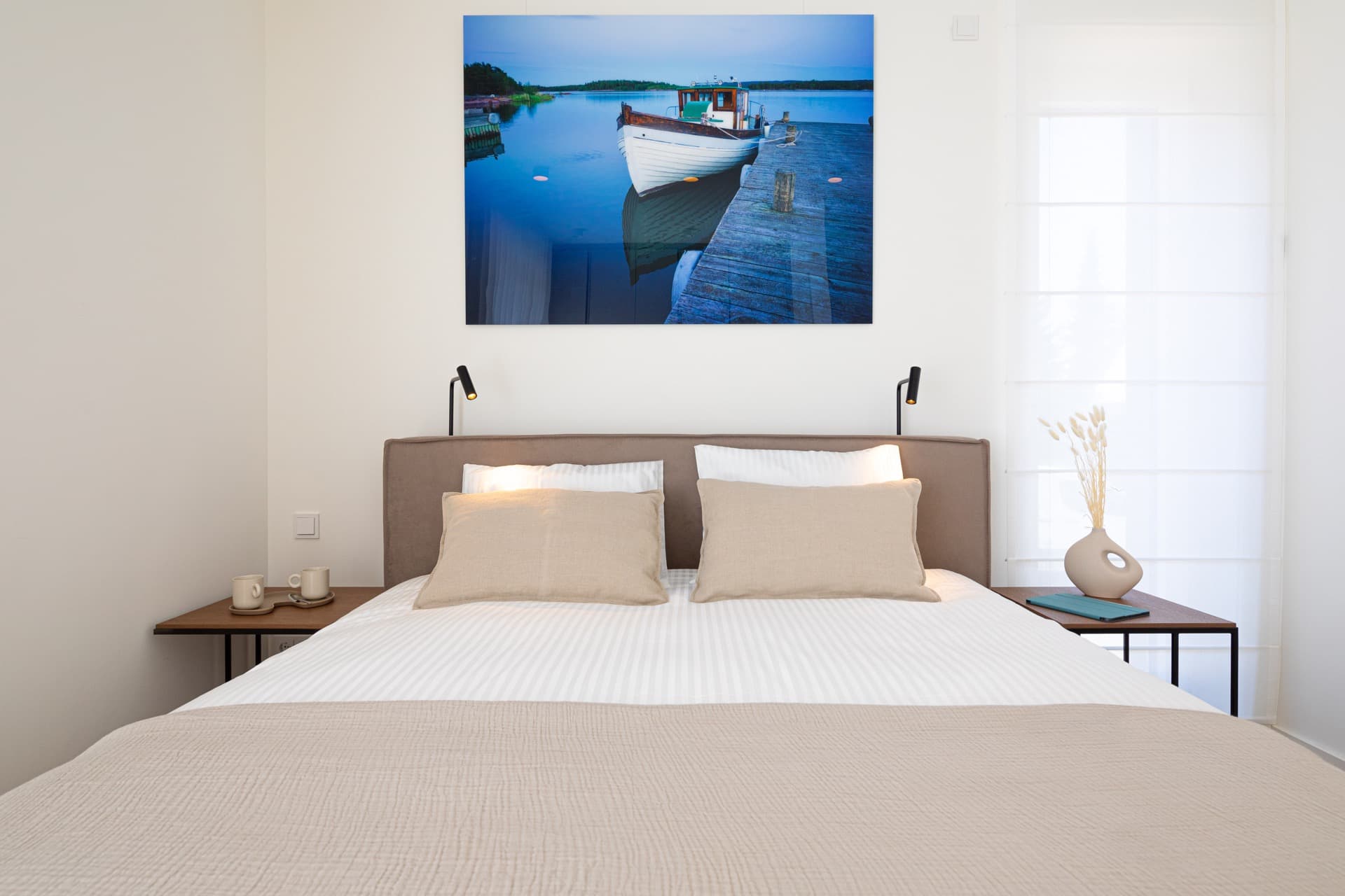 Picture of a king-size bed with white sheets, and stone-colored pillows. The bed is flanked by lamps, bedside tables with coffee cups, iPad, and dried grass vase. Large floor-to-ceiling window on the right. Above the bed, wall features a large photograph of a boat at sea.