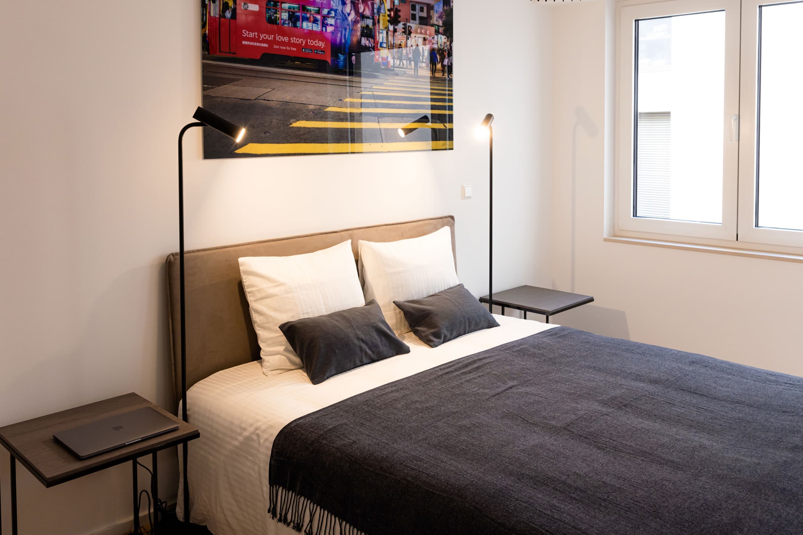 Bedroom with a large bed with white sheets and a blue cover. The bed is flanked by two nightstands and two lamps. Above the bed, hangs on the wall a big picture. On the left of the bed, there are two windows.