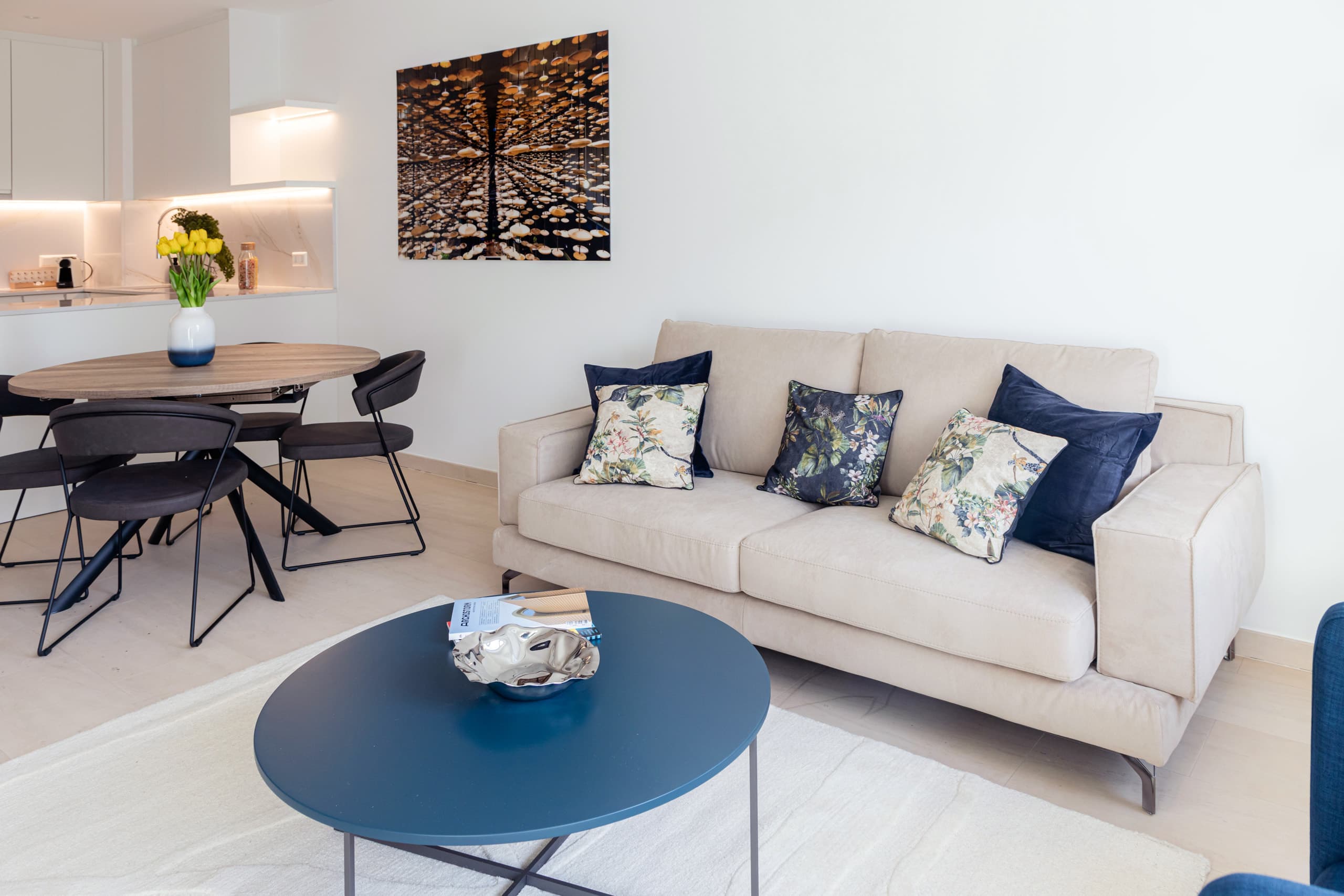 Lounge view with a sofa and cushions on it, and a coffee table in front. Underneath, a white rug. On the left, a round dining table and chairs. In the background, the kitchen can be seen.
