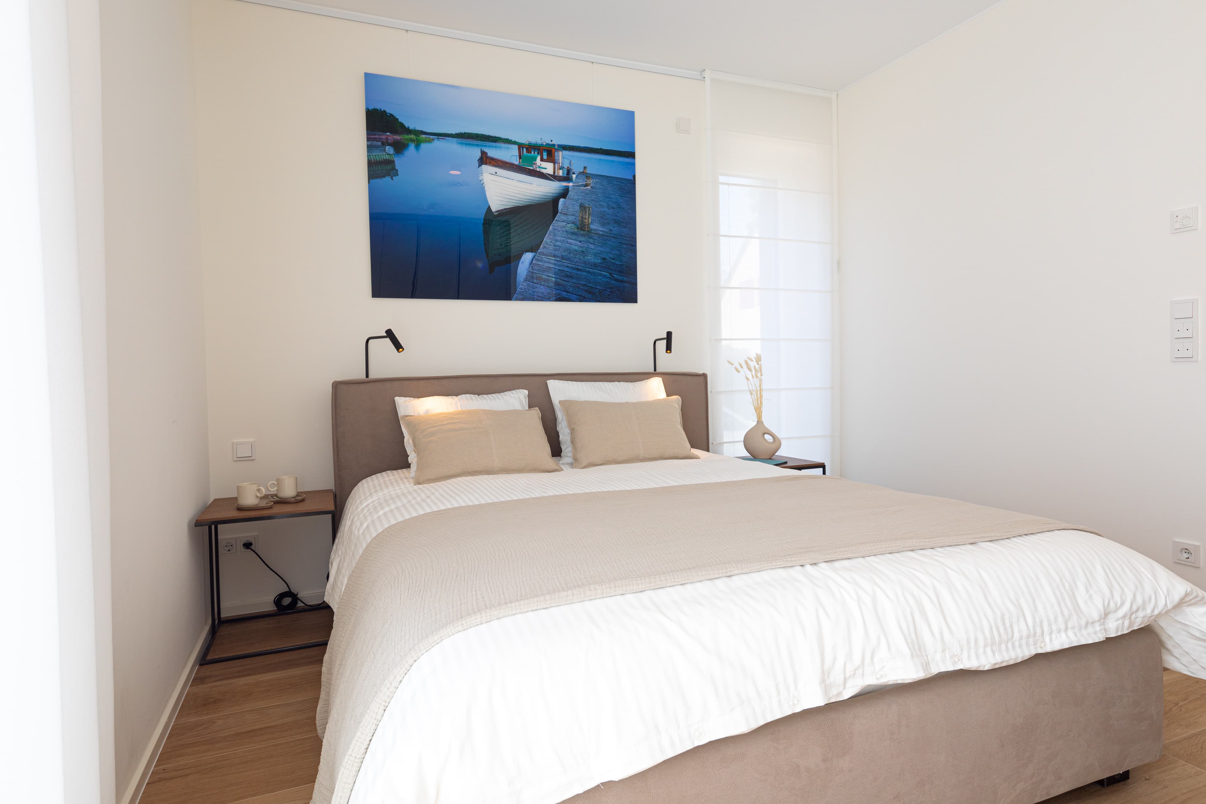 Bedroom with a large bed with white sheets and pillows, flanked by two lamps and nightstands. On the wall behind, hangs a big picture