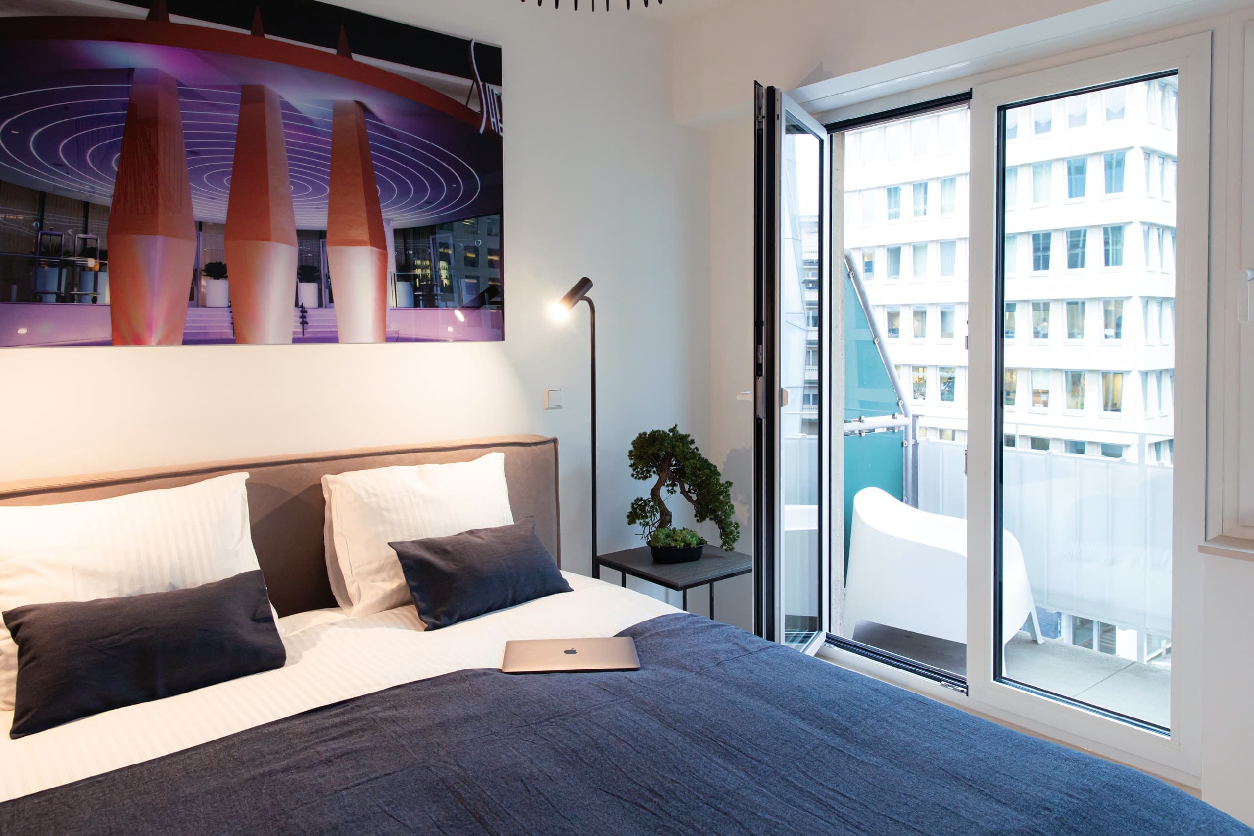 Bedroom with a large bed with white sheets and a dark blue cover. The bed is flanked by two nightstands and two lamps. Above, on the wall, hangs a big picture. On the right, there is a big balcony door and a white chair on the balcony.