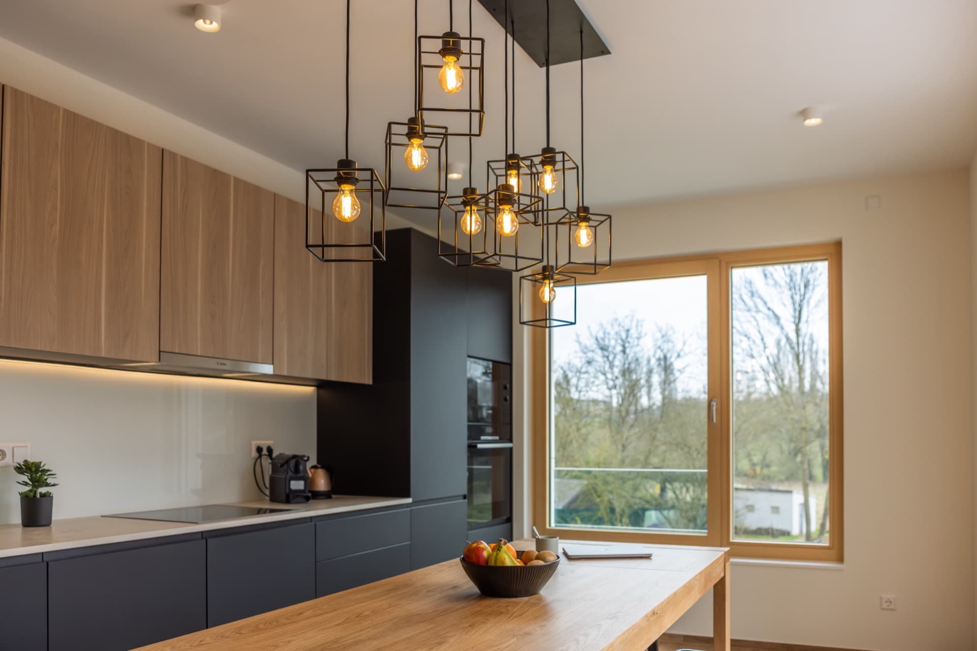 Overview over kitchen, including stylish lights, stove, oven, coffee machine, storage spaces and tall table.