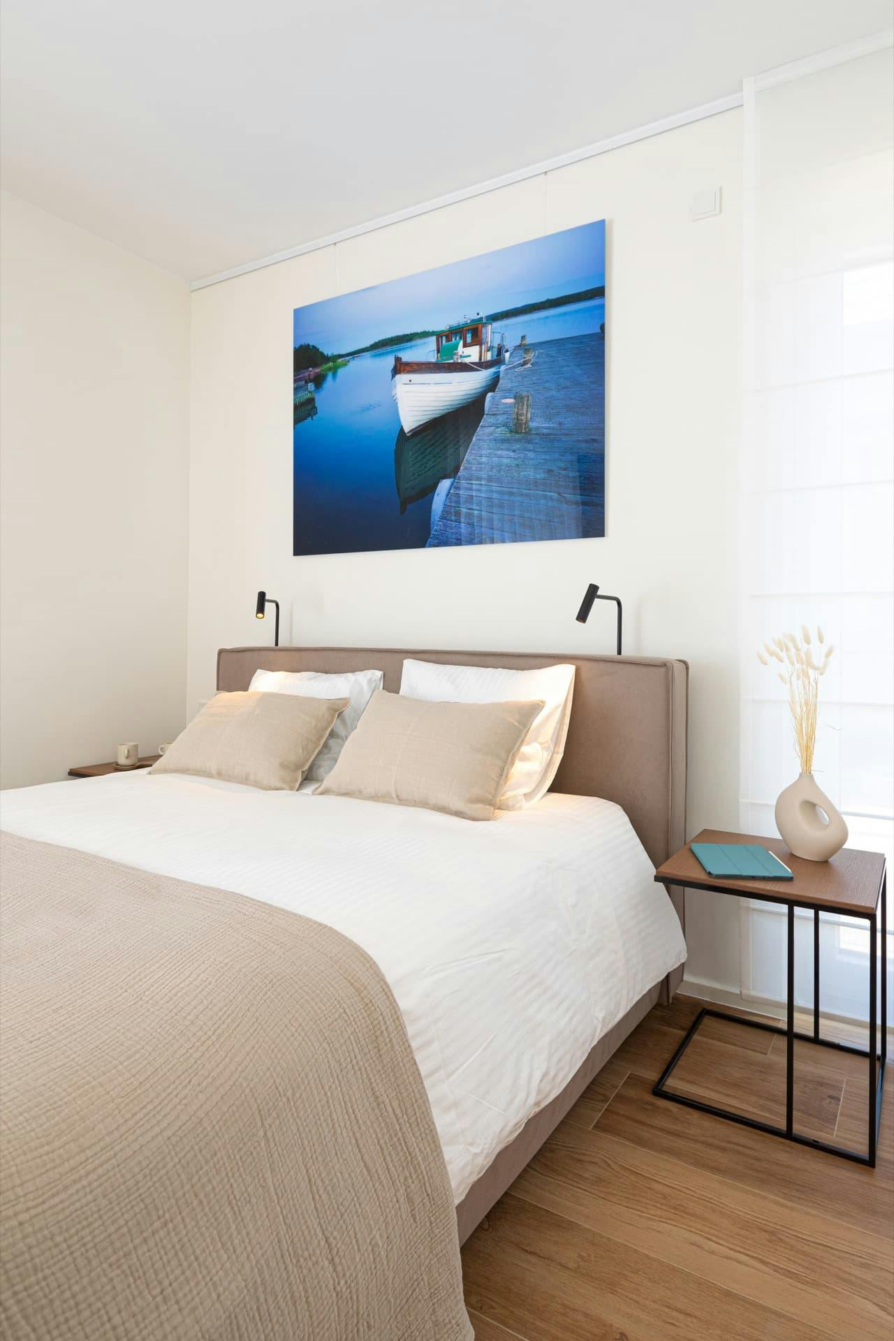 a bed with white bedding and stone coloured pillows with bedside table and a large floor to ceiling window on the right. There are coffee cups, an ipad and a vase of dried grasses on the tables and a large photograph of a boat at sea above the bed.