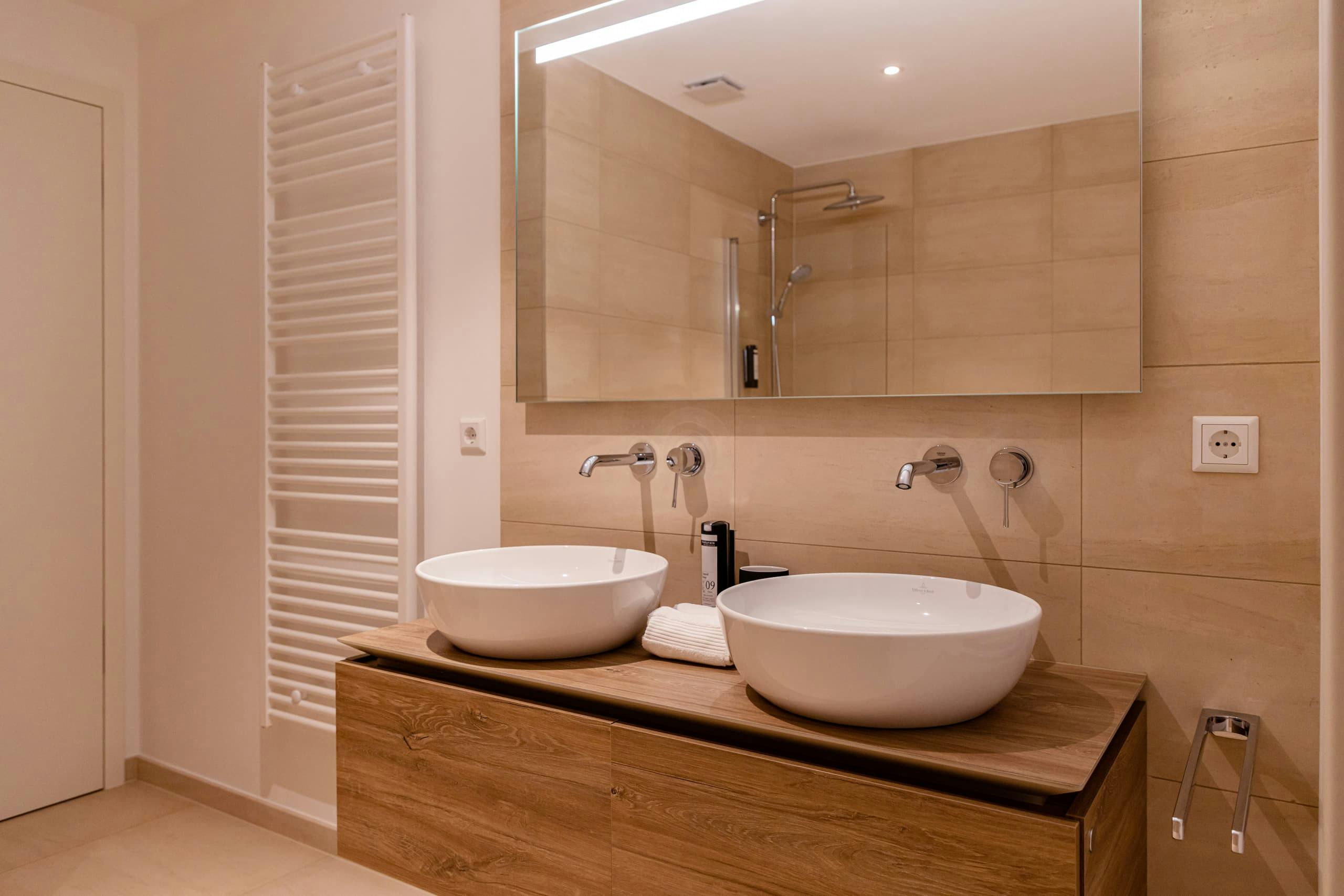Bathroom with two sinks, one next to the other, and a heating radiator on the left. Above the sinks there is a mirror, and on its right, an electricity plug and towels hanger.