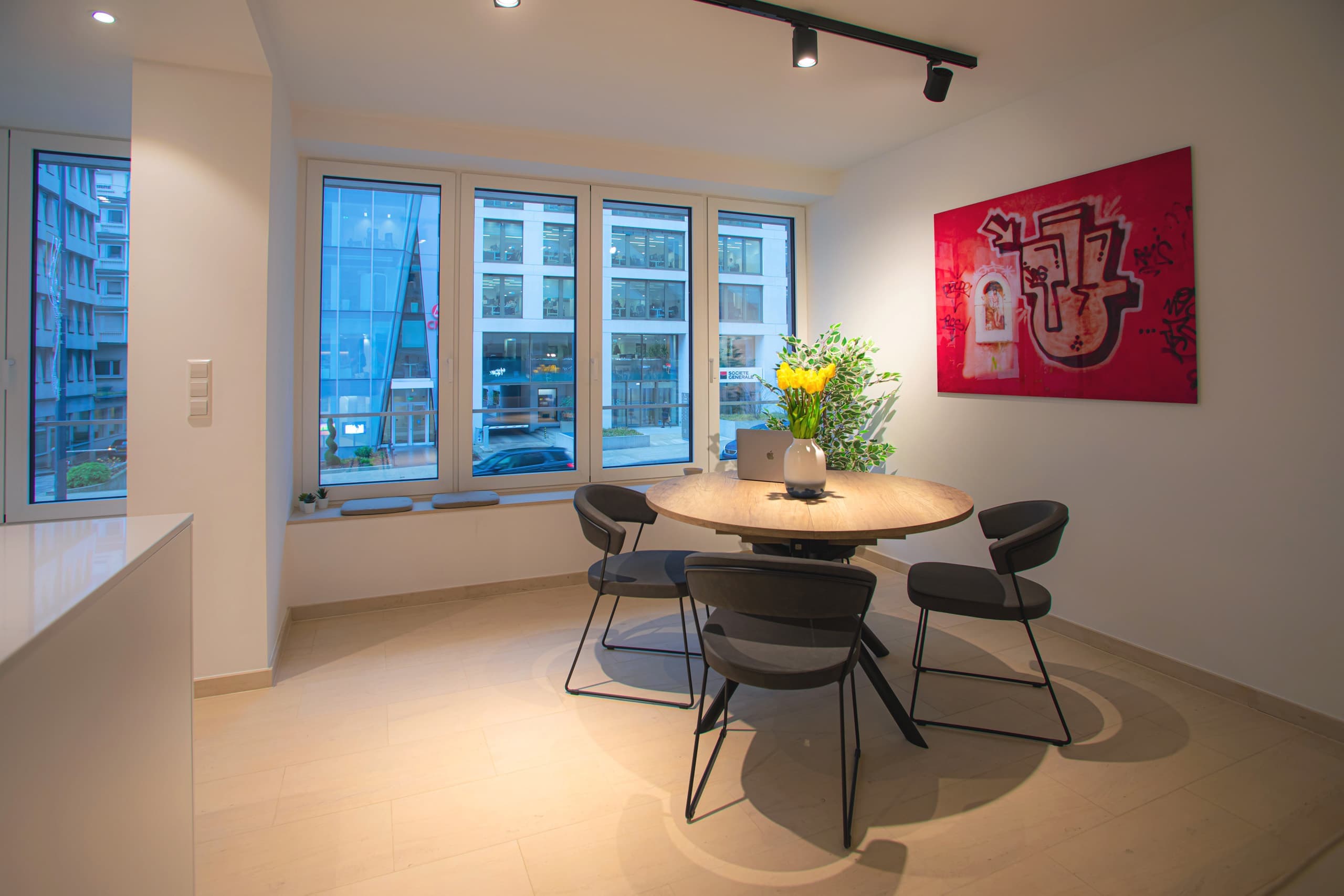 Round dining table with chairs, behind it there are large windows and a big flower pot. On the wall, on the right, hangs a big picture. On the left, the corner of the countertop can be seen