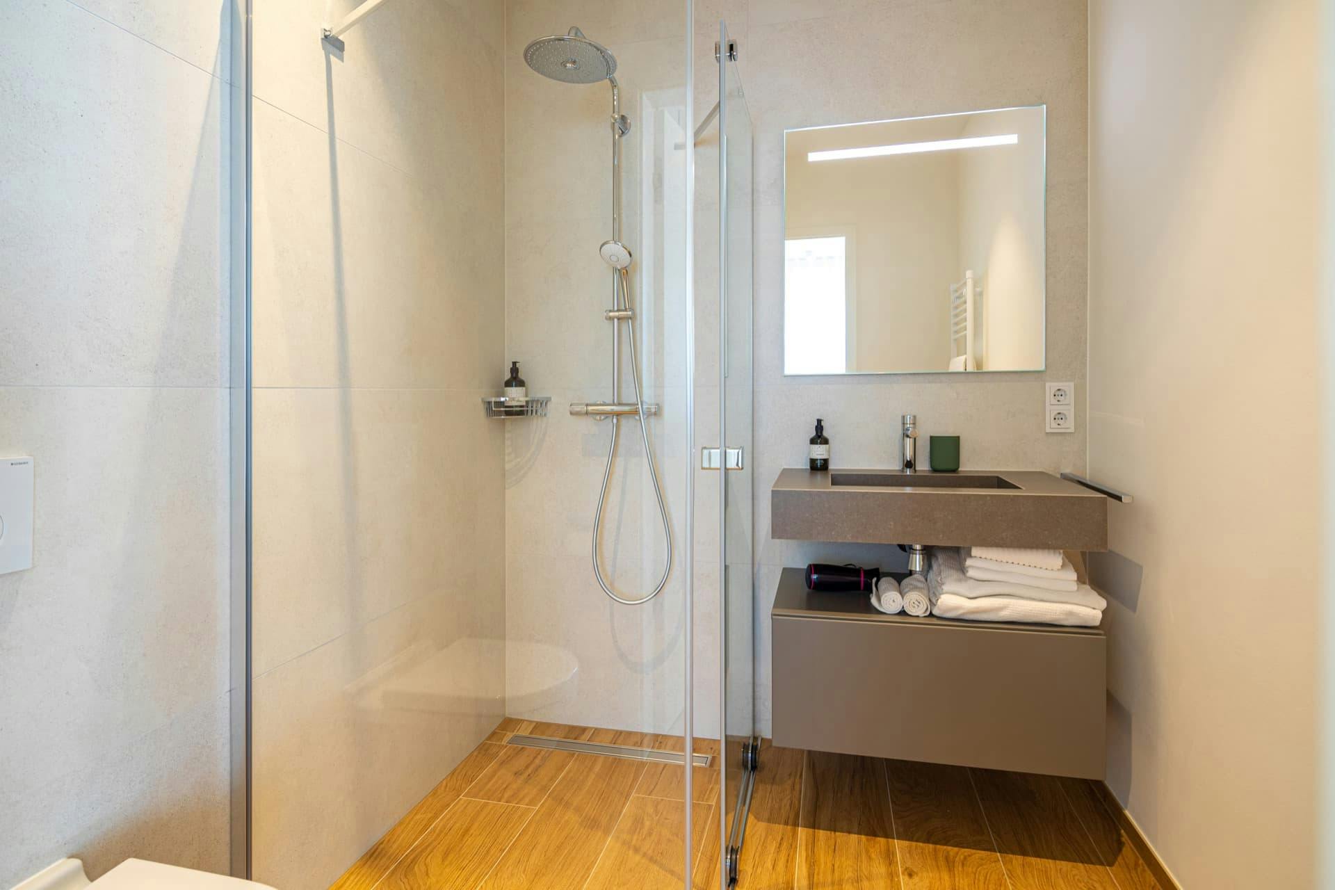 En-suite bathroom picture displaying a shower with shower gel, a sink with soap, a toothbrush cup, and towels below. Reflected heat radiator in the mirror above the sink, with two electric sockets on the right.