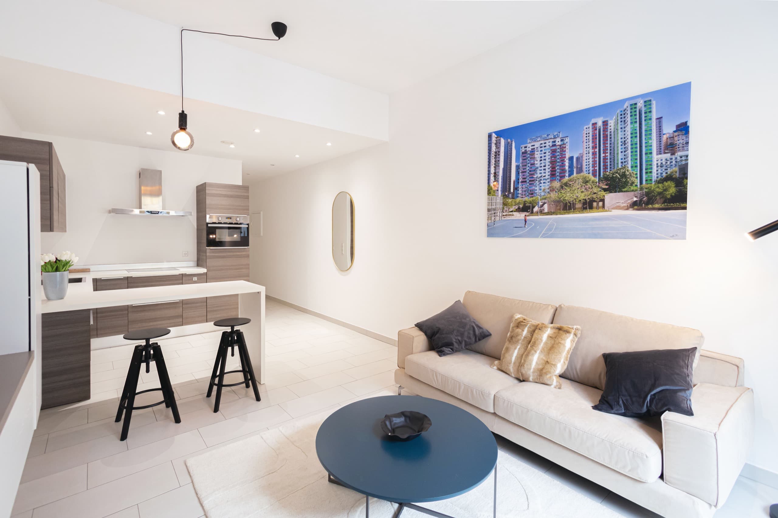 Open-plan lounge and fully equipped kitchen, with tall countertop and chairs. Sofa with cushions, coffee table on a white carpet. A big picture hangs on the wall behind the sofa.