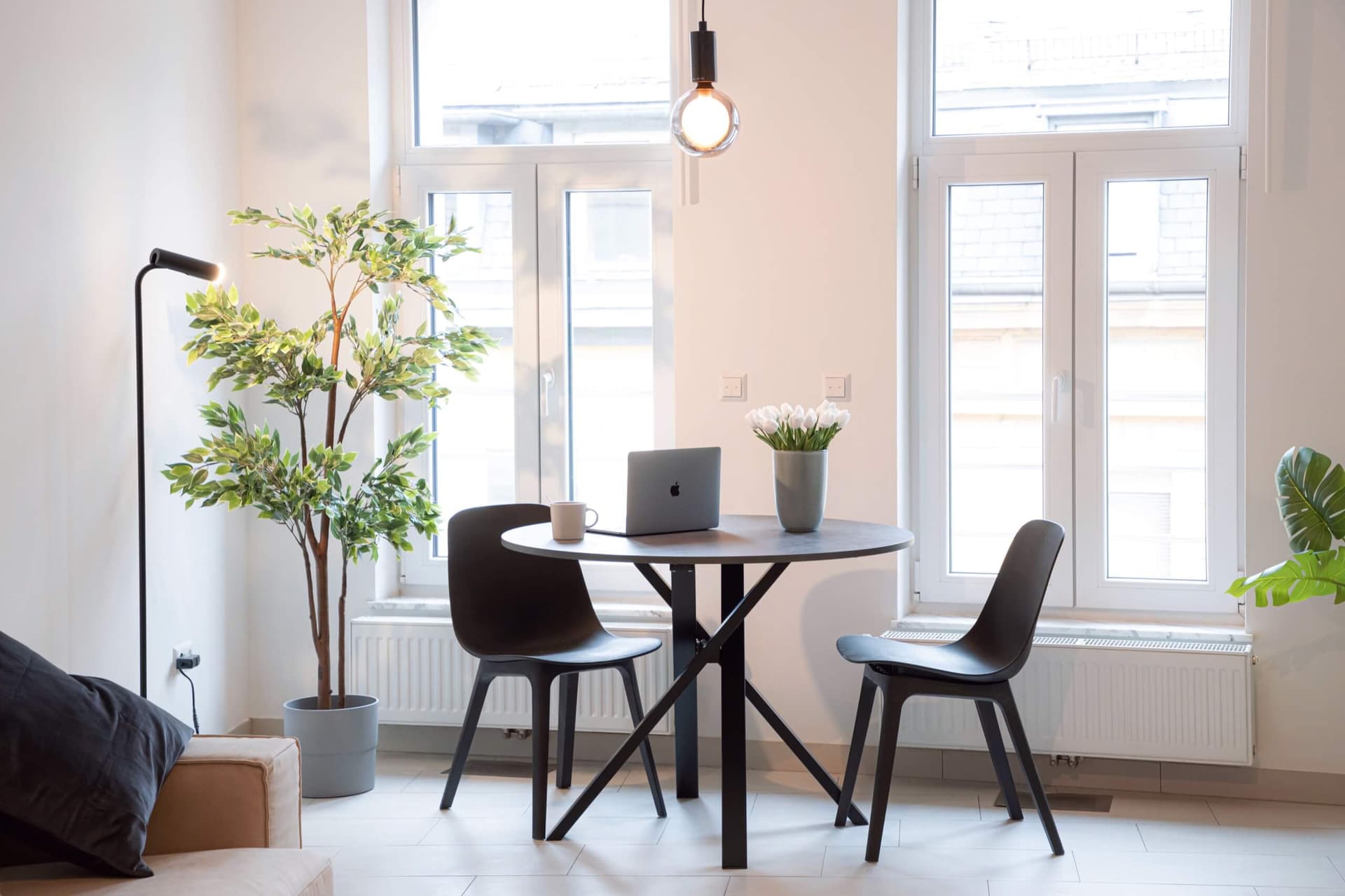 The inside of an apartment looking towards the 2 windows.  In front there is a table with 2 chairs. Sitting on top of the table is a macbook pro, a vase with white tulips and a coffee cup.  Next to the window is a large green plant.
