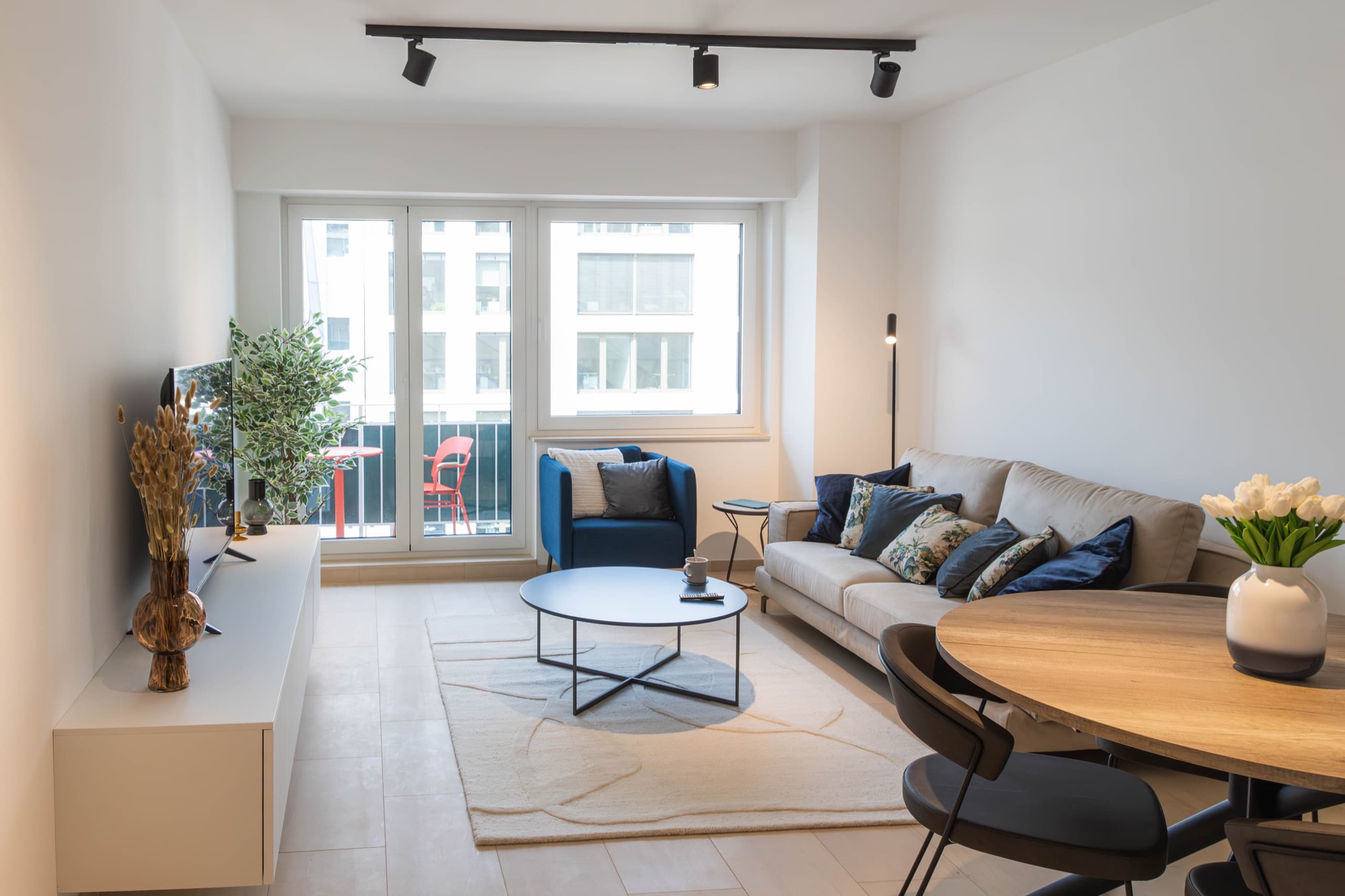 Lounge area with a TV stand with a smart TV on it, on the left. On the right, there is the dining table with chairs and a flower vase on top. Next to it, there is a couch with cushions. In front of the couch, there is a coffee table with a white rug underneath. In front of the windows and balcony door, there is an armchair with a small coffee table next.
