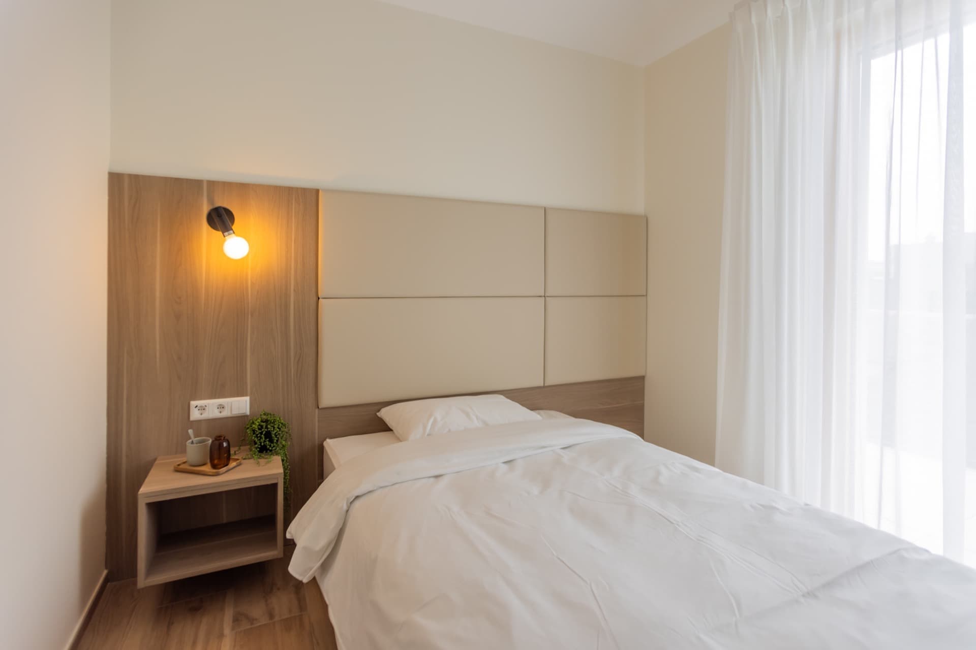 Overview of the bedroom. In the center is a bed with high-quality white linens. On its left are a nightstand, a lamp, and light switchers. On its right is a large floor-to-ceiling window.