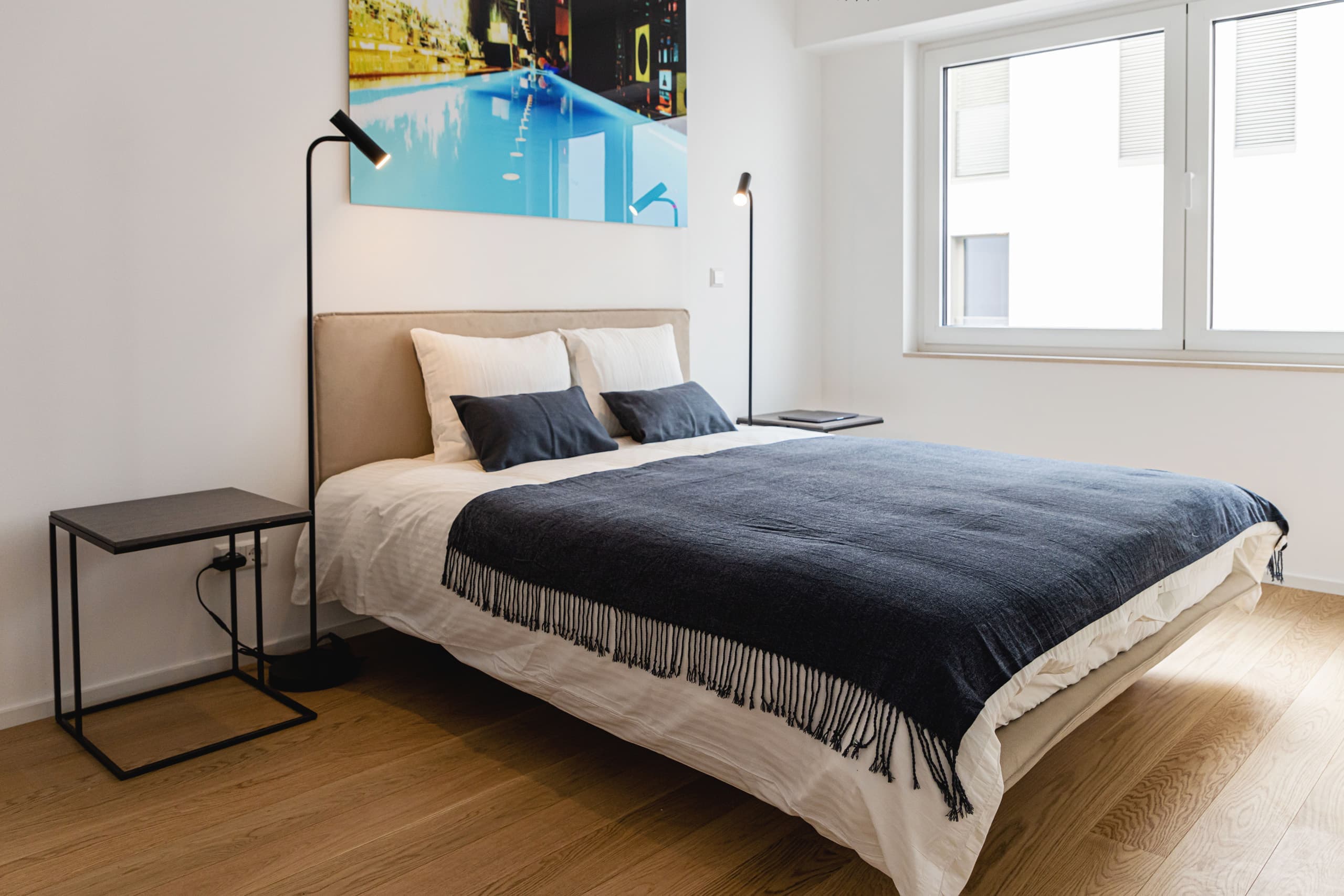 Bedroom with one large bed flanked by two standing lamps and two nightstands. Above the bed hangs a big picture, and on its right, there are two large windows.