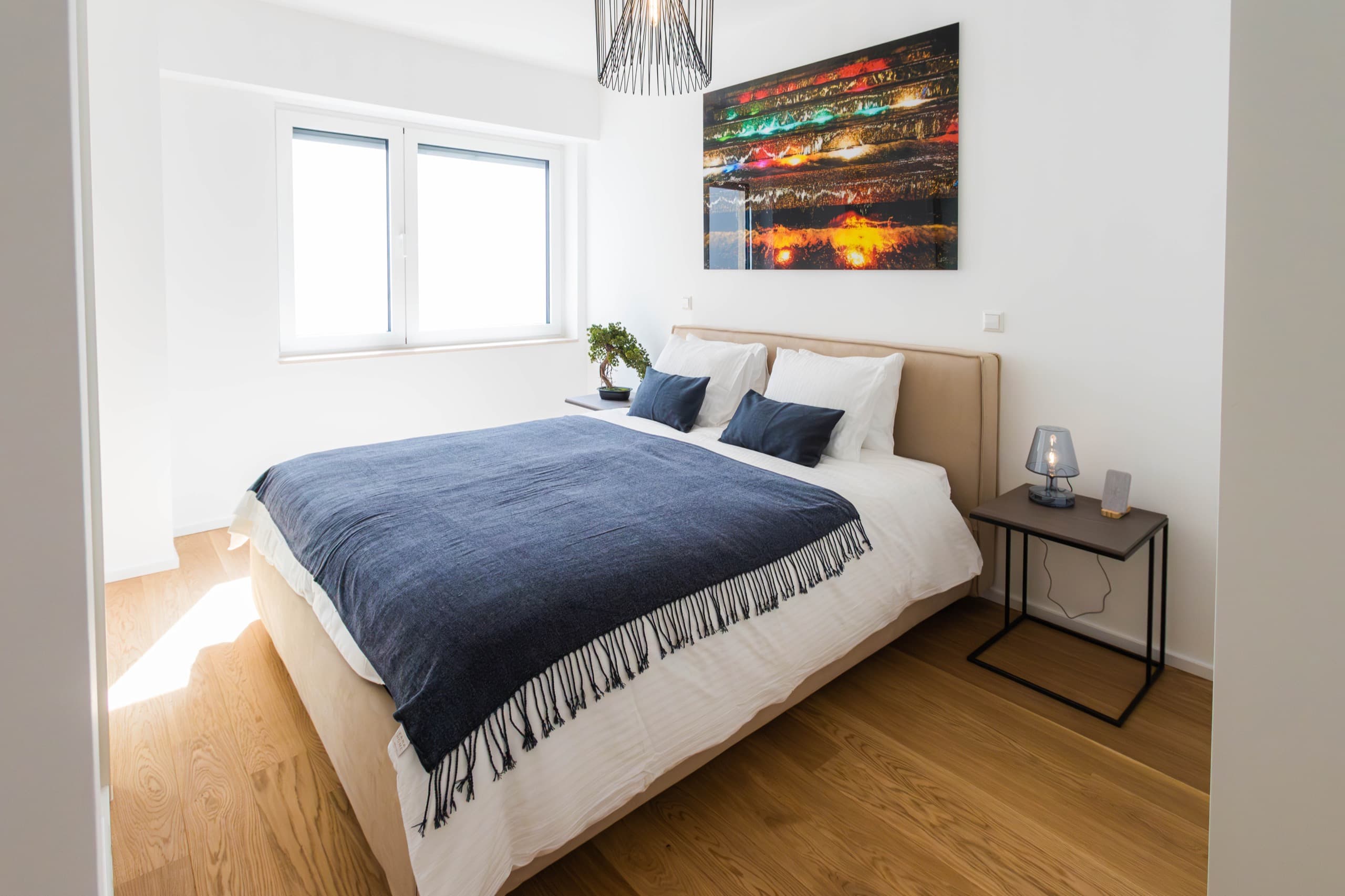 Bedroom with a large bed with white sheets and a dark blue cover on it. The bed is flanked by two nightstands. Large windows, on the right of the bed, light up the room. Above, on the wall, hangs a big picture.
