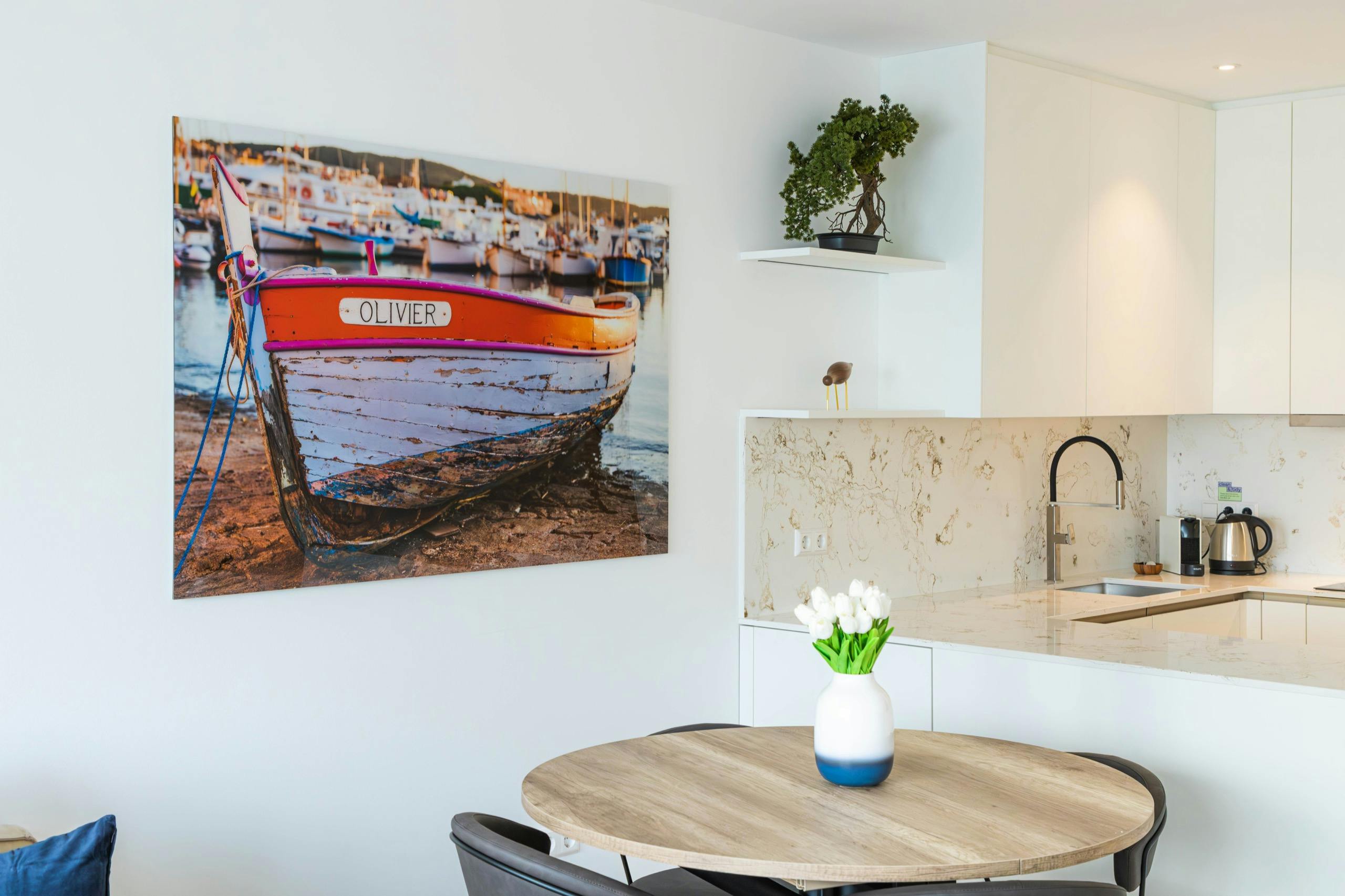 Kitchen and dining area zone comprised of a round dining table with chairs and a flower vase on top. On the wall hangs a big picture with a boat ashore.