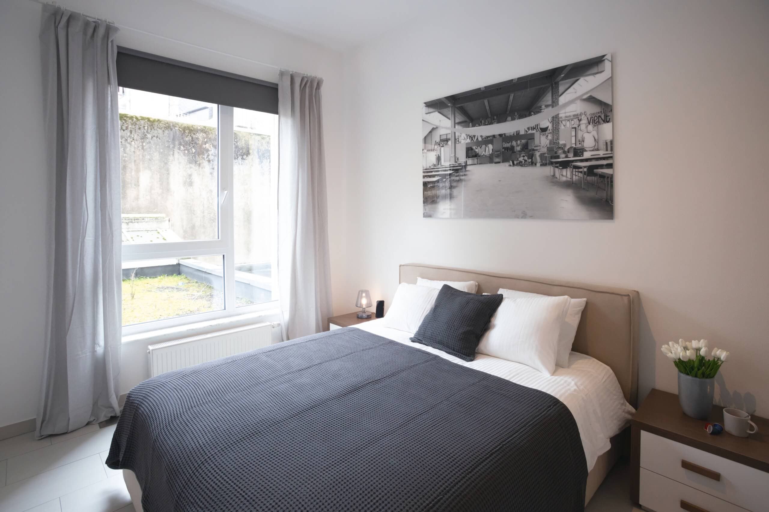 Bedroom with large window drapes with light grey curtains. A bed with blue cover over white bedding. There is a transparent lamp and wireless phone charger next to the bed.