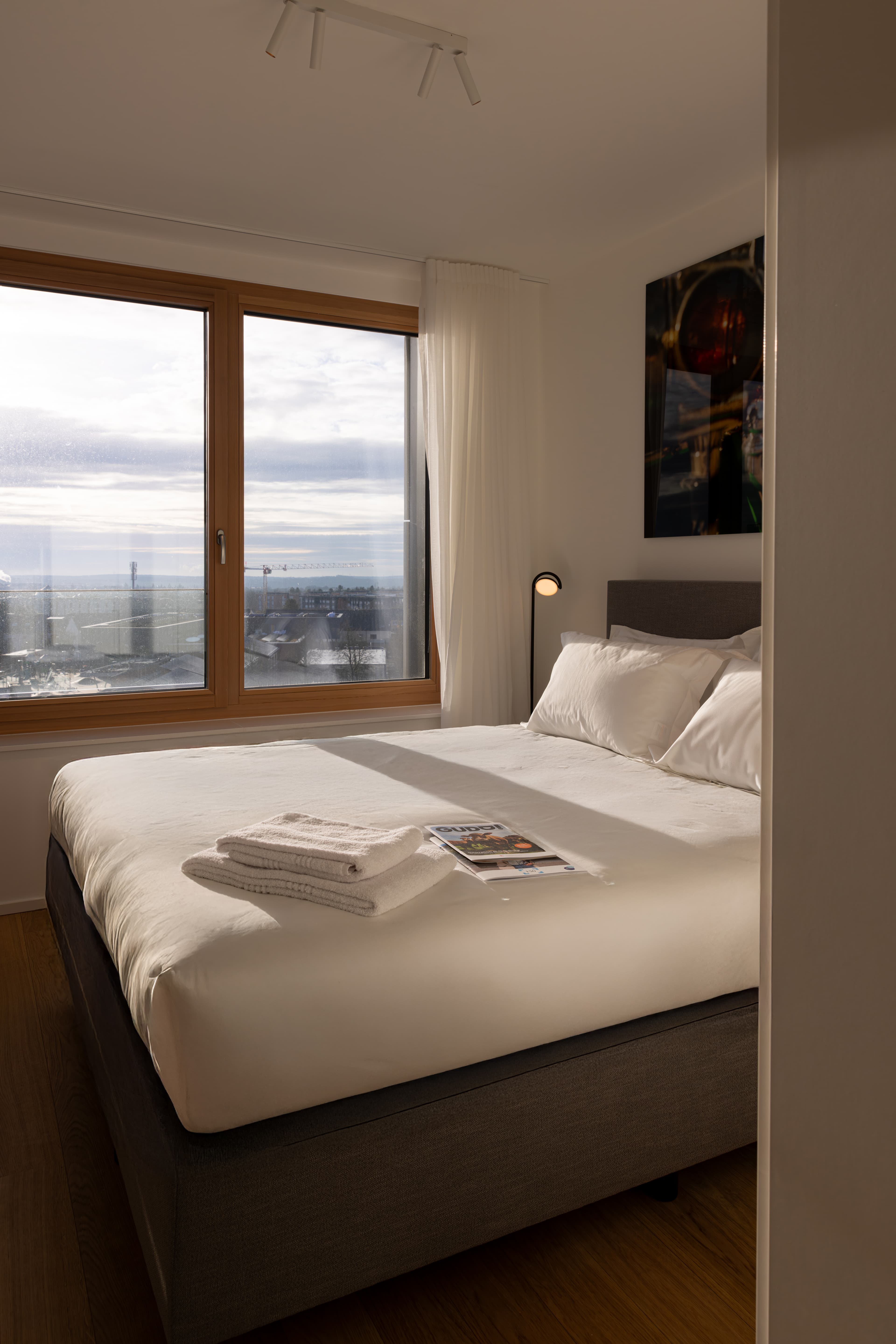 Bedroom with king-size bed and high-quality linens. On the right side of the bed there is a night stand with a lamp on it and, also, one large window.
Above the bed hangs a large framed photo