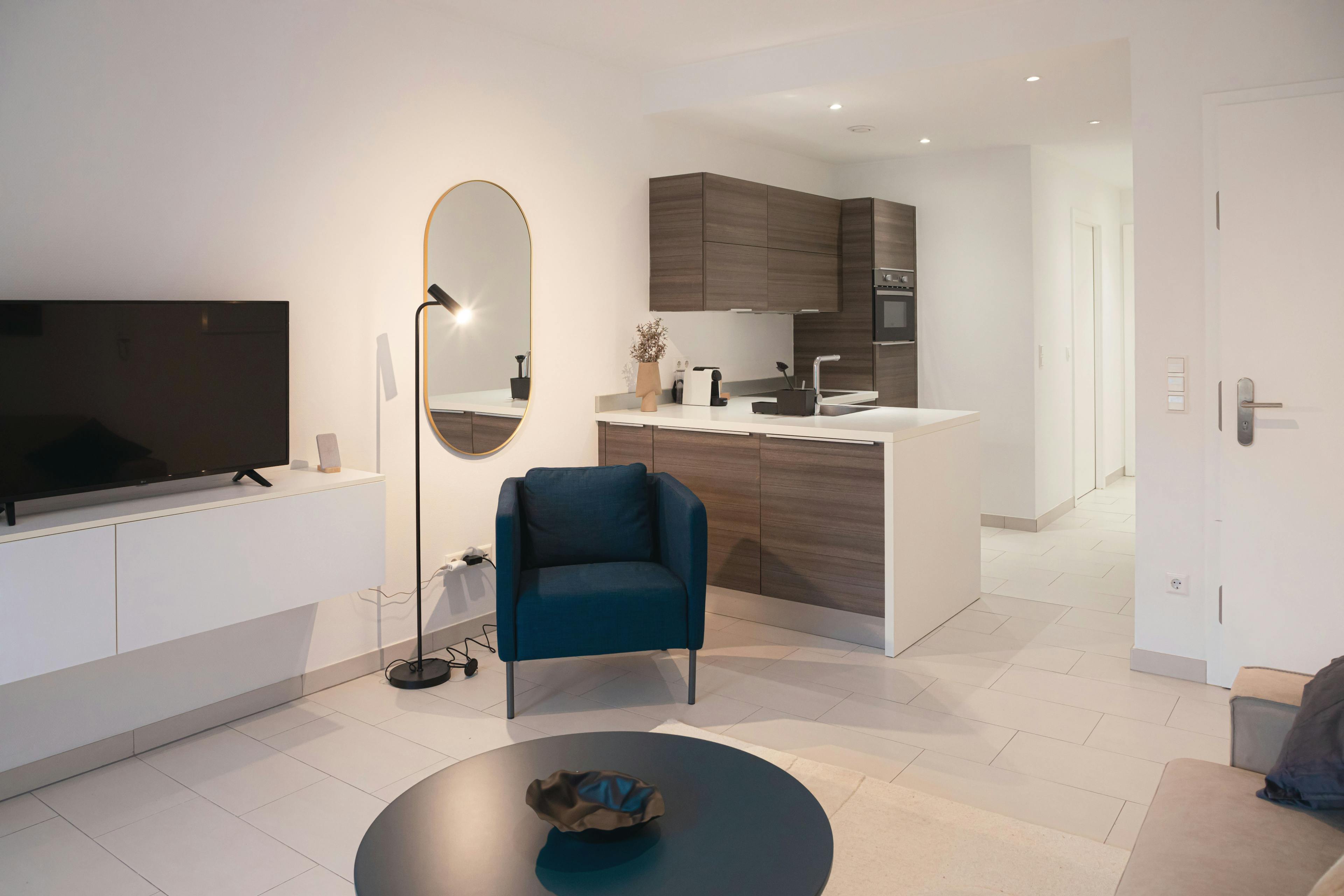 Zoomed-out image of the lounge and kitchen area, including the hallways in the background. It contains a  TV stand with a smart TV on it, an armchair next to a standing lamp, and a mirror on the wall. In front of the hallway, there is the fully equipped kitchen.