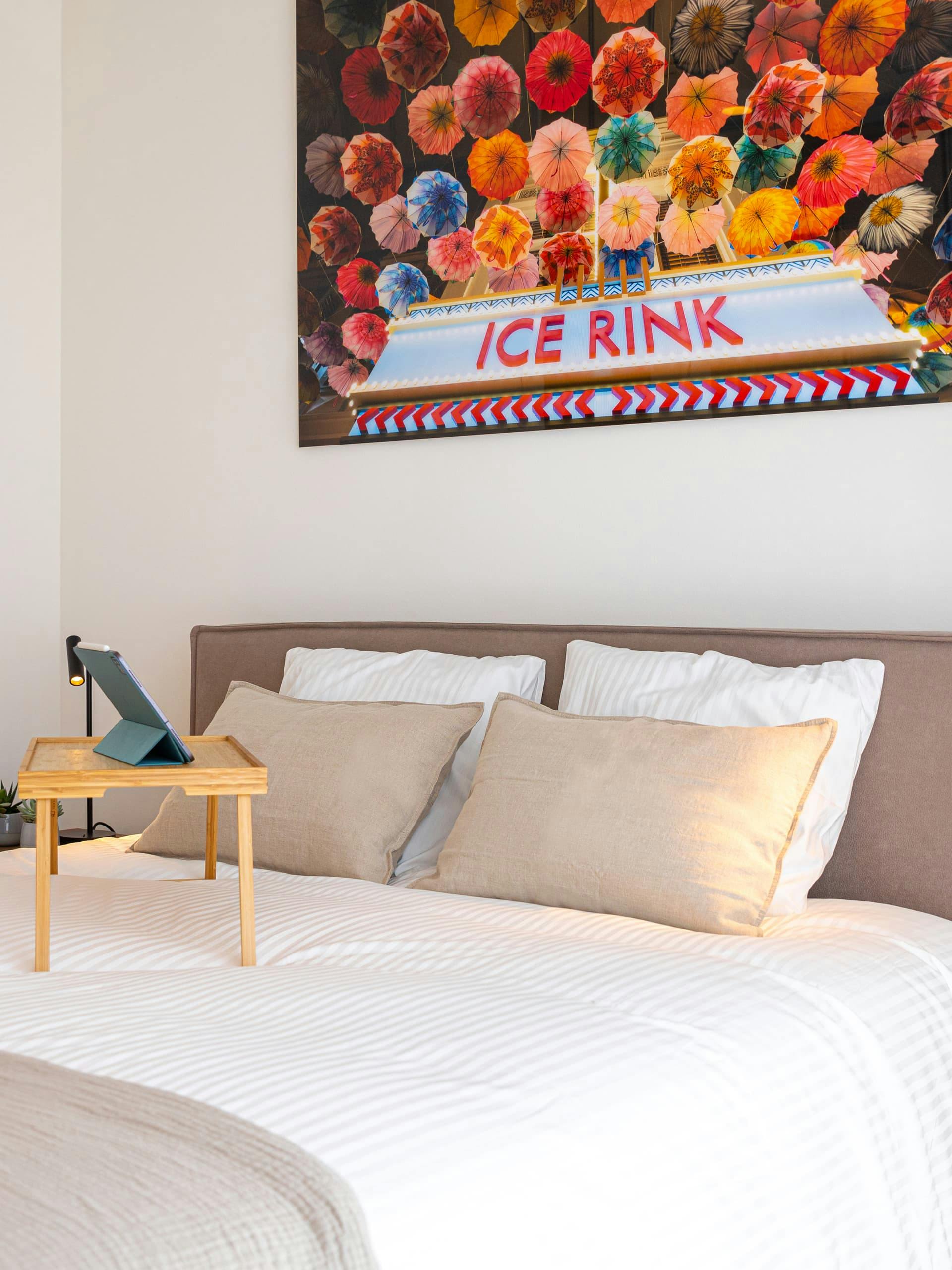 View of a bed, sitting on top there is a tray with legs and an ipad on its stand. above the bed there is a photograph of flowers and the words Ice Rink.