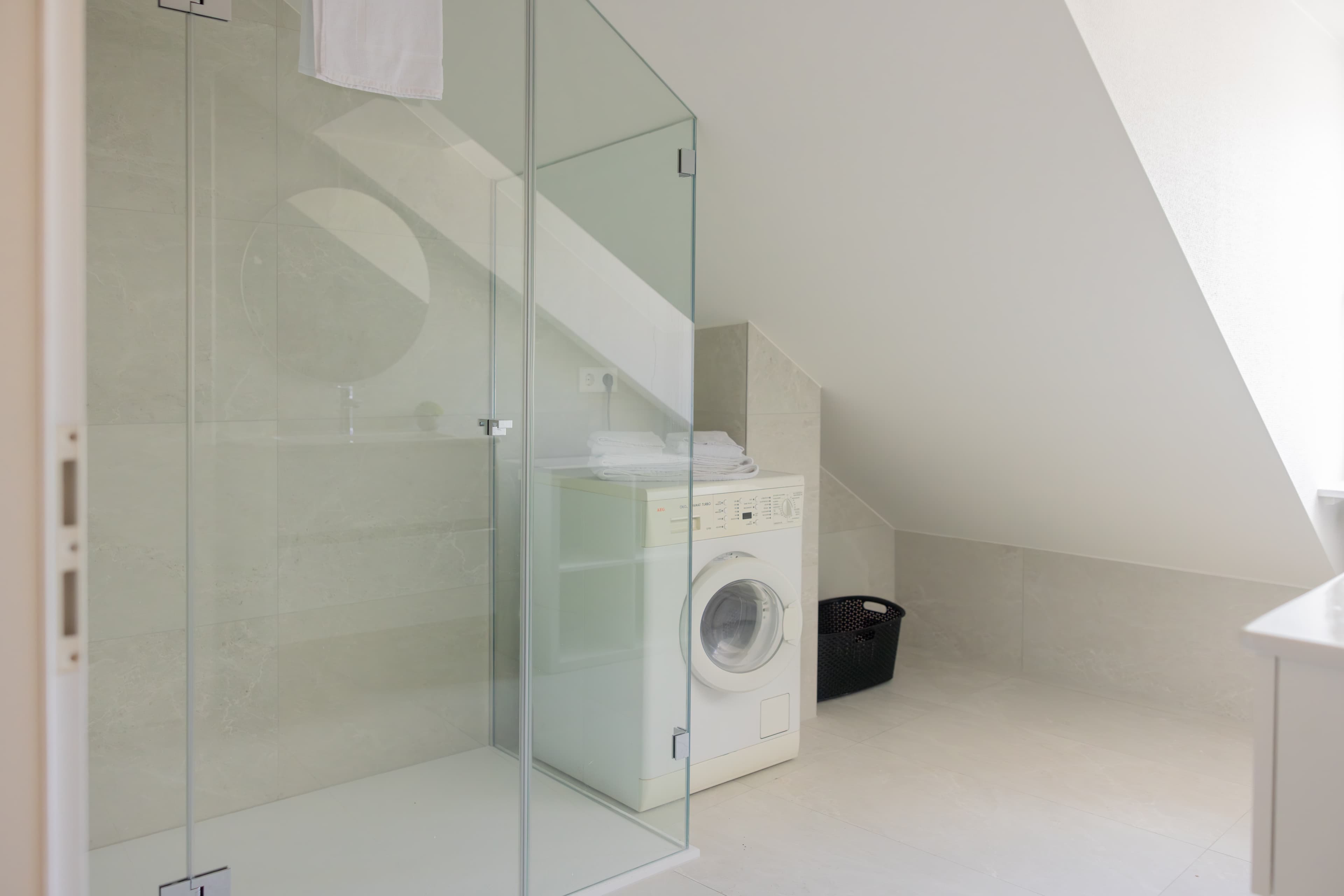 The picture depicts a washing machine and a laundry basket. There is also a walk-in shower cabin, a sink, and a mirror