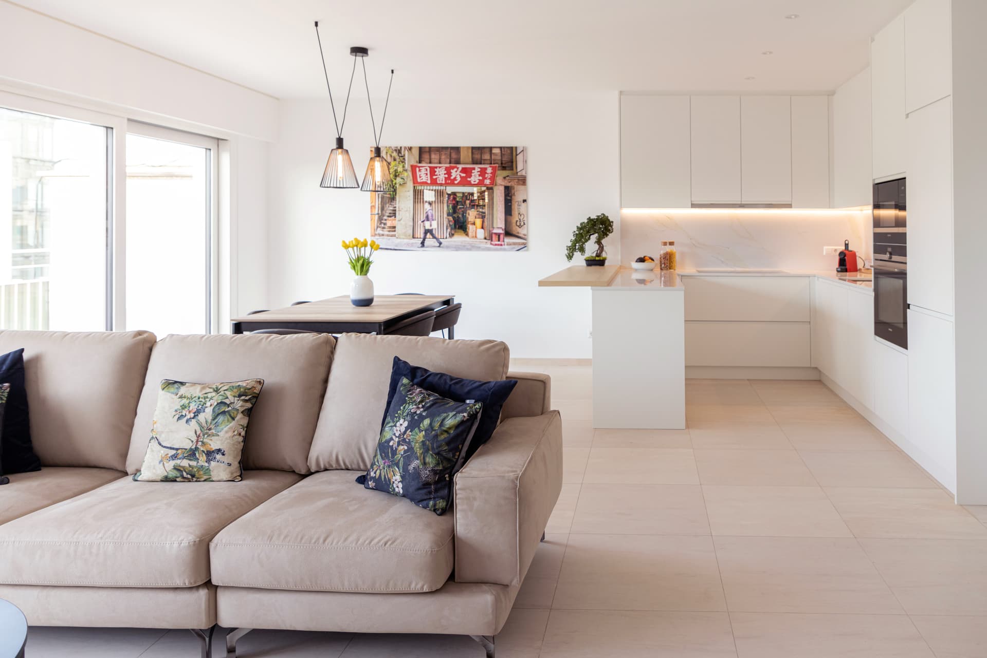 In the background, a table with a flower vase on it, and a picture of a street in hong kong hanging on the wall. Next, on the right, is the kitchen with a bonsai flower pot on a counter. In the foreground, is a beige sofa with multi-color cushions on it.