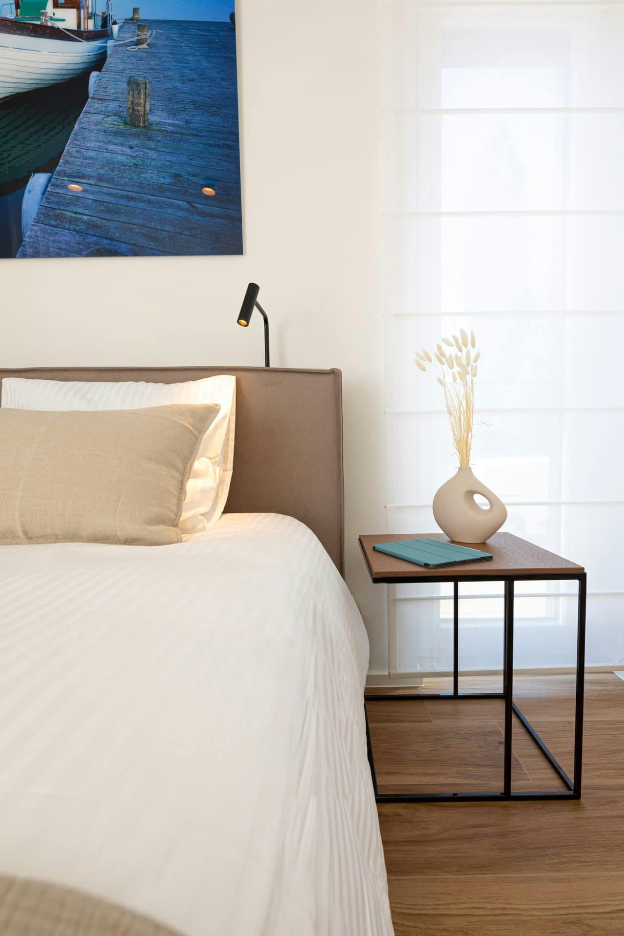 image shows a small part of a bed with a bedside table and a large full length window. on the table there is an ipad with a blue cover and a vase with a handle holding dried grasses