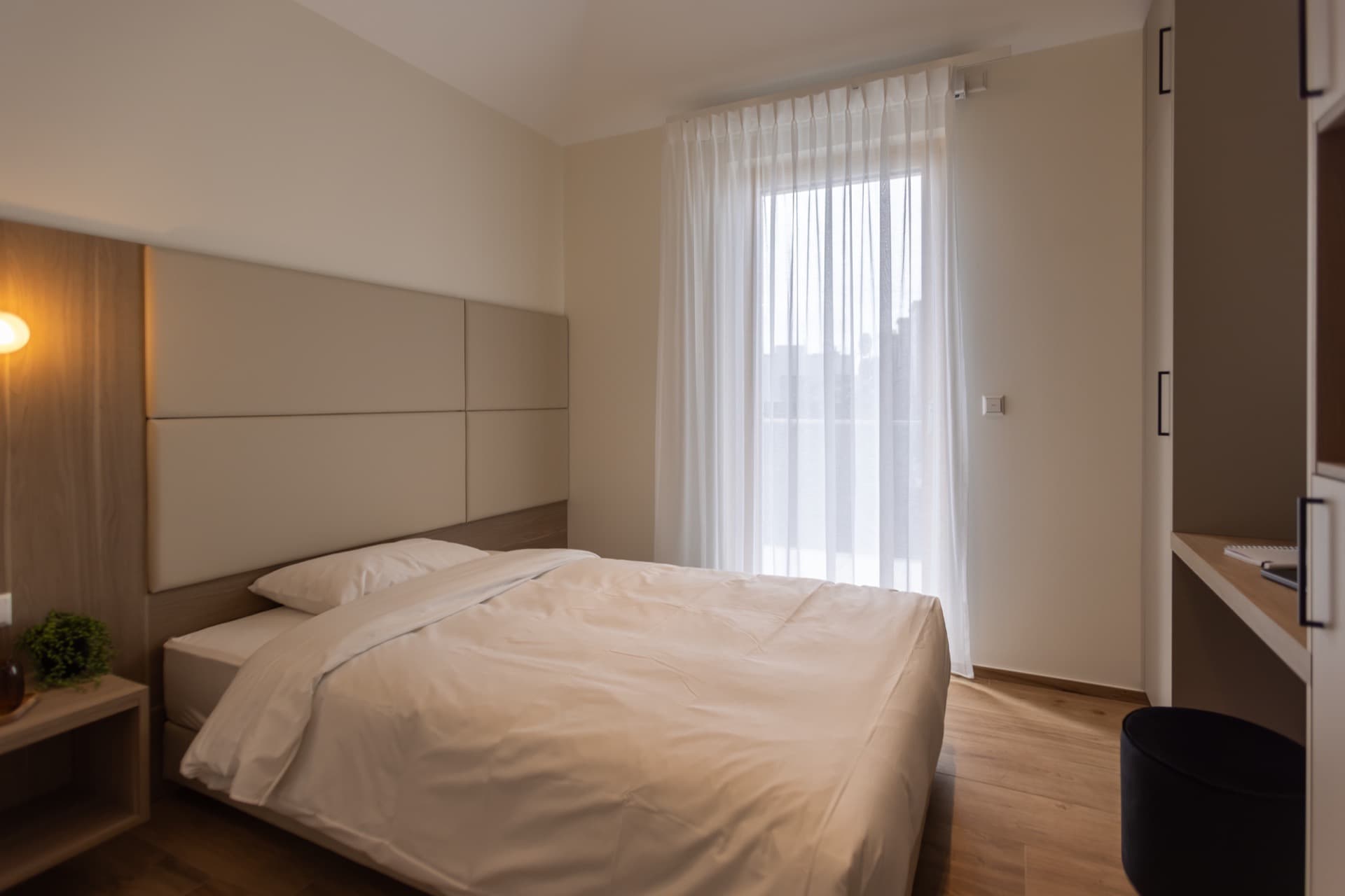 Overview of the bedroom, having a framed double-bed with high-quality linen. Next to it there is a large floor-to-ceiling window with curtains. 
In front of the bed there is a working desk.