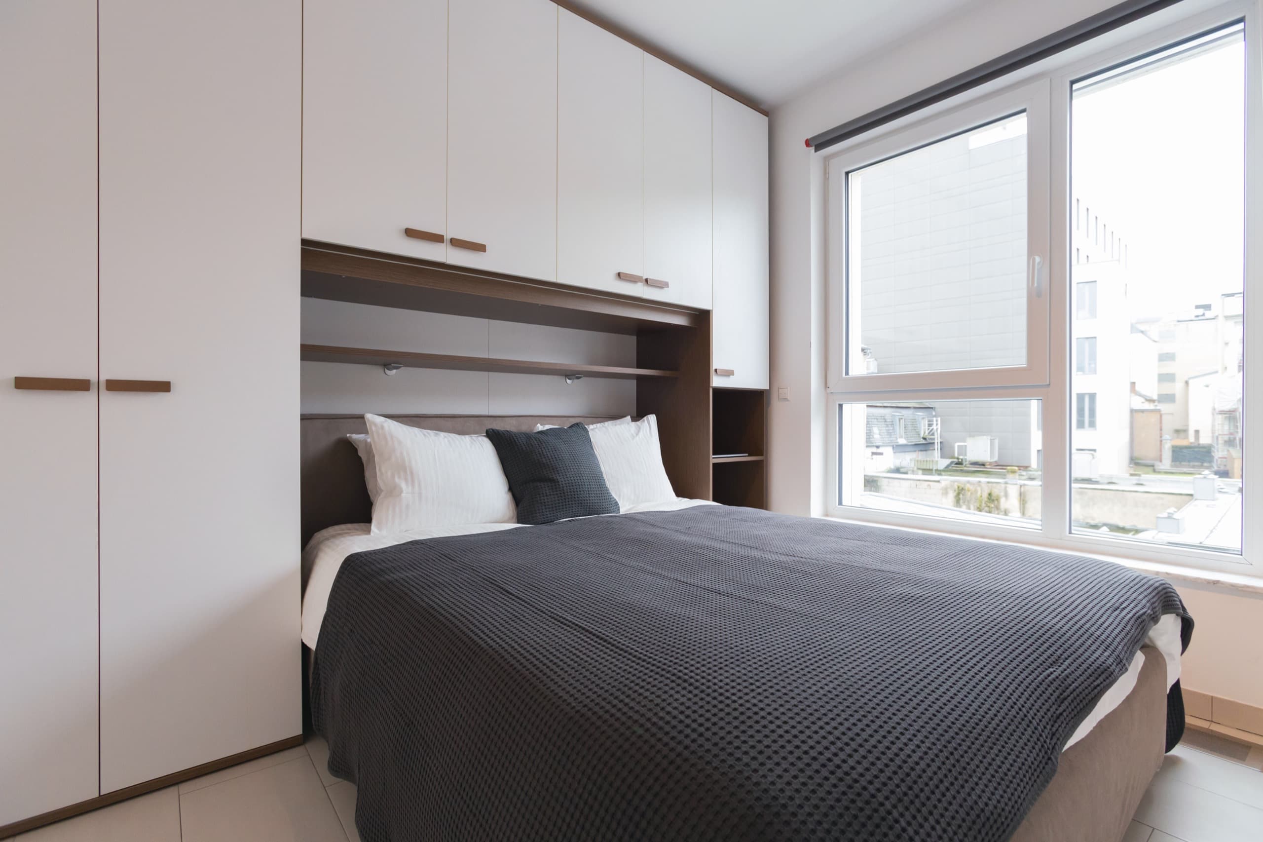 Bedroom with a large bed under the under-measure dressing. The bed has white sheets and cushions and a dark blue cover. On its right, is a large window
