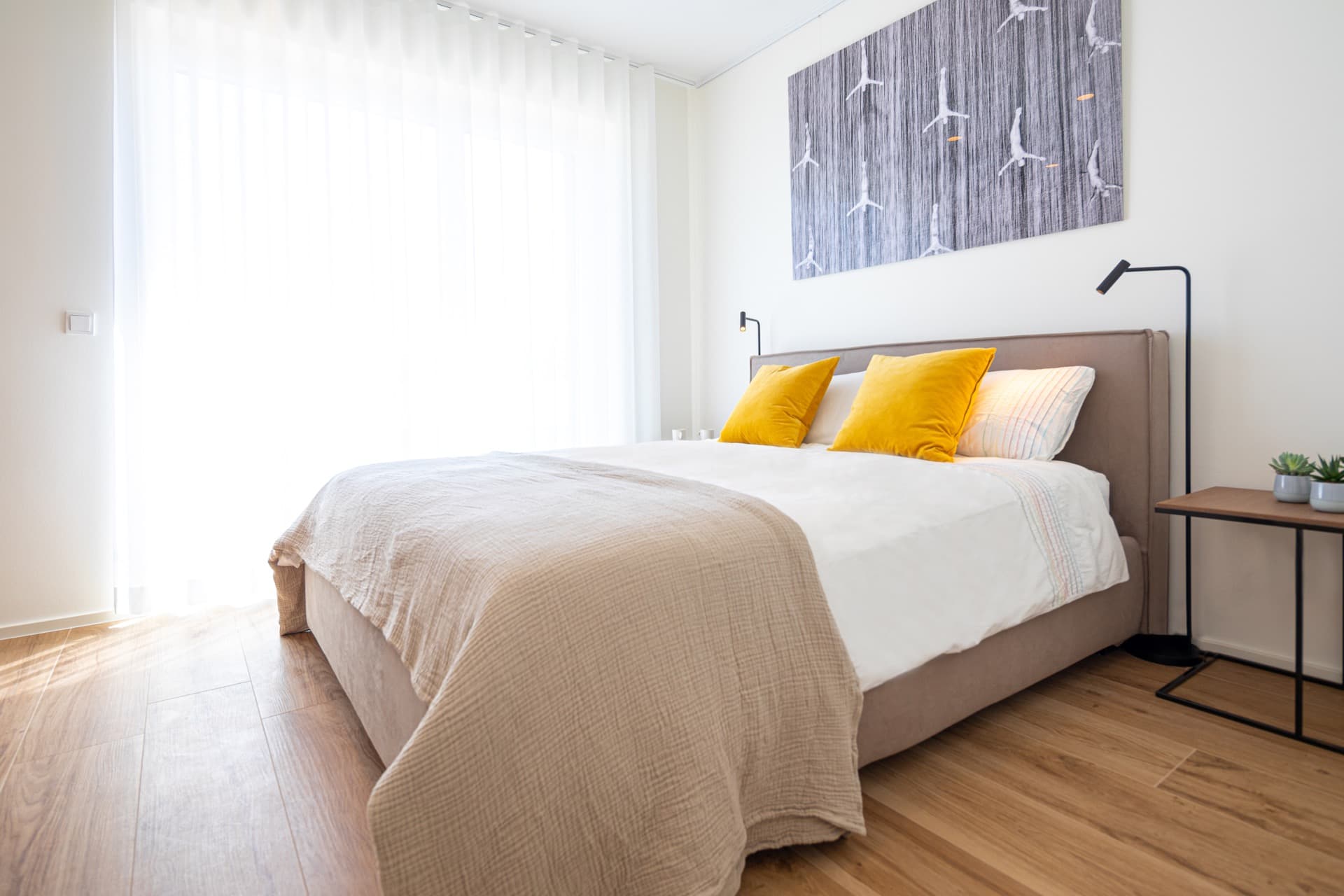 The picture depicts a bed with white sheets and mustard cushions, with two lamps flanking it. Hanging on the wall, there is a picture. Behind there is a large floor to ceiling window with sheer curtains.