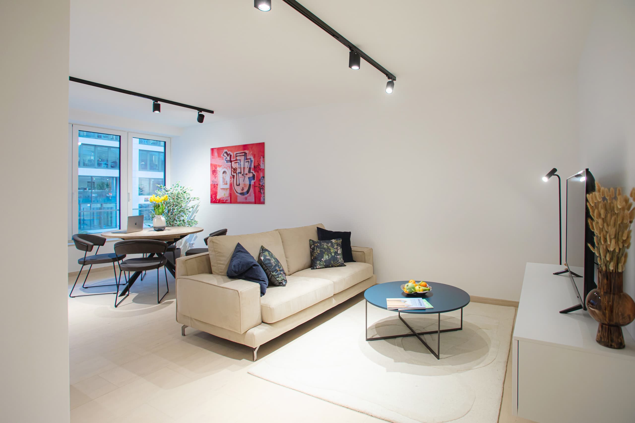 Lounge image with a round dining table next to the large windows, behind is a couch with cushions and a coffee table on a white carpet. In front of the couch is a TV stand with a smart TV on it. 
On the wall, next to the windows, hangs a big picture.