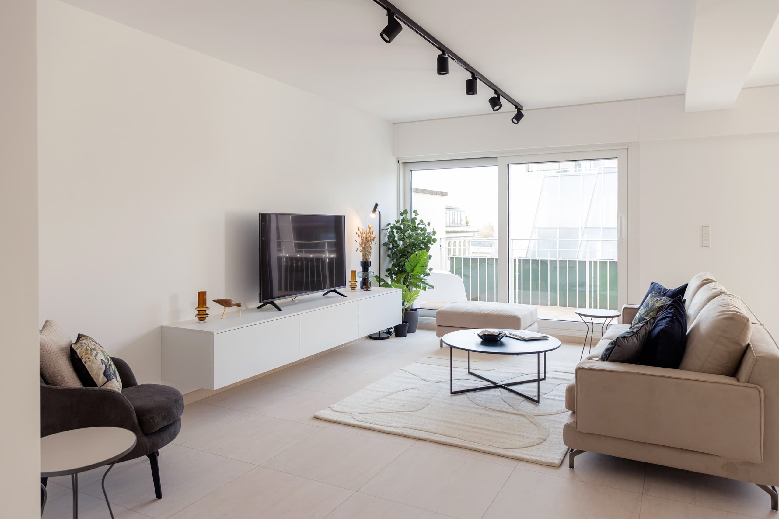 On the left, an armchair and a small coffee table. In front of it, a TV stand with a smart TV, a lamp and big flower pots. In the centre, a medium-sized coffee table, a beige carpet, and another armchair. On the right, a sofa with cushions.