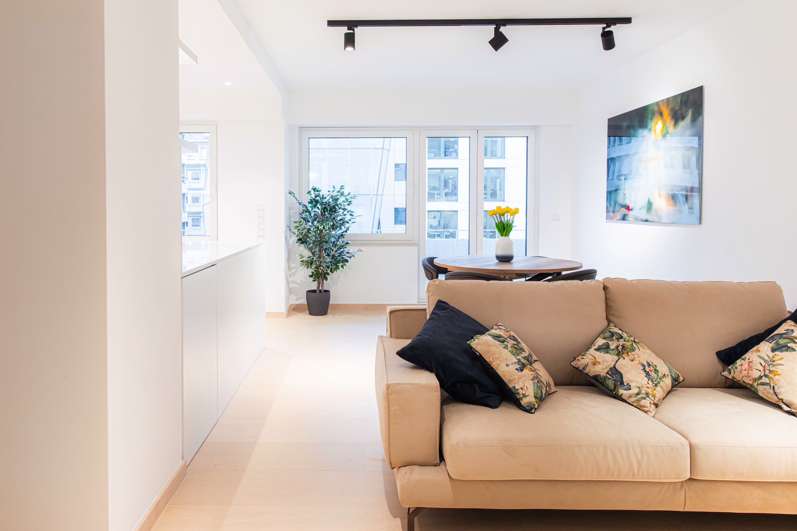 Sofa with cushions on it. On the left, the corner of the kitchen counter can be seen. Behind the sofa is a round dining table with chairs and a flower vase on it. Behind, are large windows. On the wall, next to the table, hangs a big picture